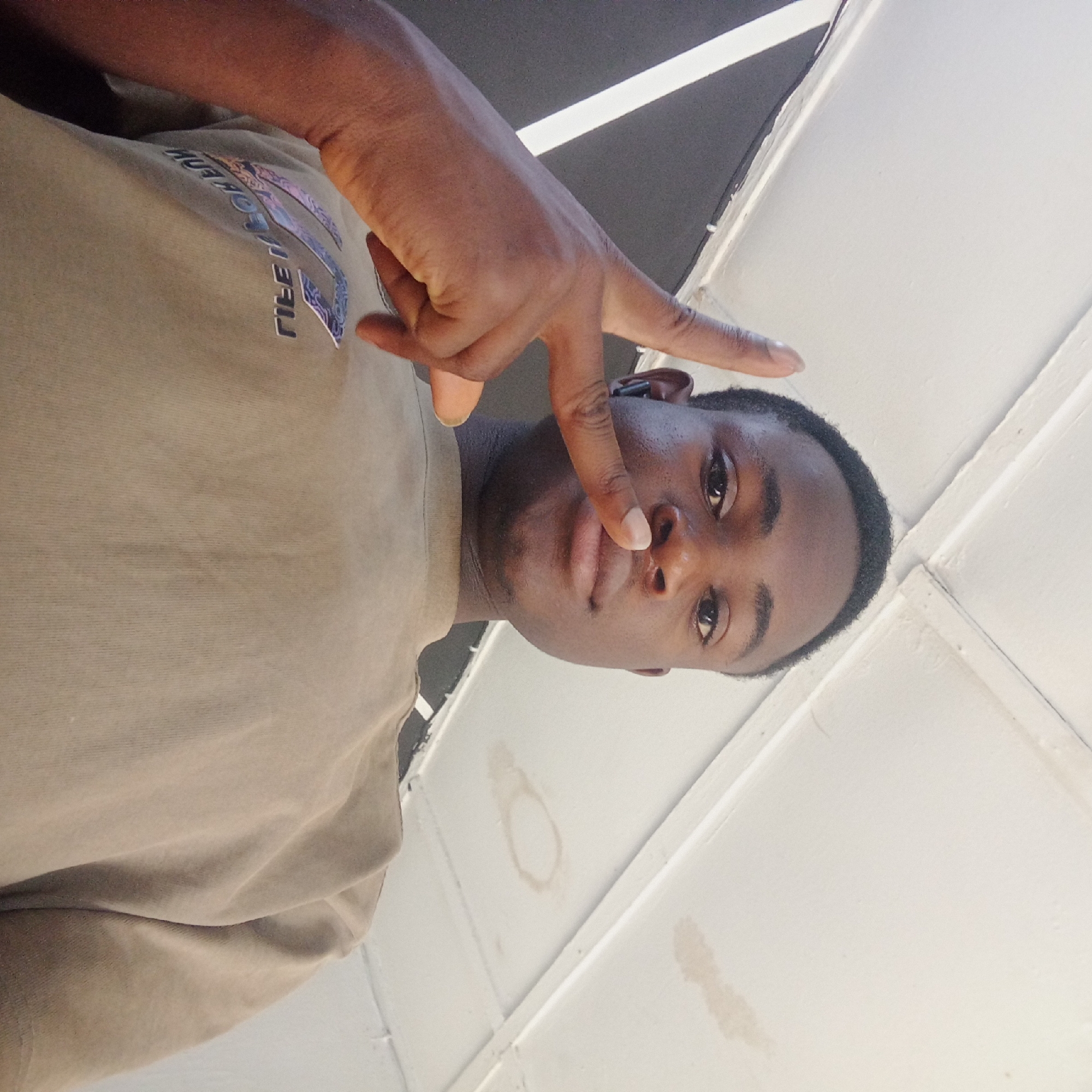 A young Black man is depicted in a close-up, low-angle shot, looking directly at the camera. He is wearing a light-colored, possibly beige or light brown, short-sleeved t-shirt. His right hand is raised, forming a 'V' gesture with his extended index and middle fingers, while the other fingers are bent. The tip of his index finger is positioned near his nose.
On the front of his t-shirt, visible in mirrored form due to the image's orientation, are two lines of reflective or holographic text. The upper line clearly reads "AUTHORISED." Below this, the second line, featuring stylized letters, appears to spell "PIECES," with the initial 'P' rendered similarly to the Greek letter Pi (Π).
The background consists of a white ceiling or overhead structure, featuring visible seams or panels. Several light brown, circular, and irregular stains, consistent with water damage or dirt, are noticeable on the white surface. A dark area in the upper left likely indicates a structural beam or shadow. The lighting is bright and evenly diffused, suggesting an indoor setting during the daytime. The man's facial expression is neutral. This scene is located in Jos, Nigeria.