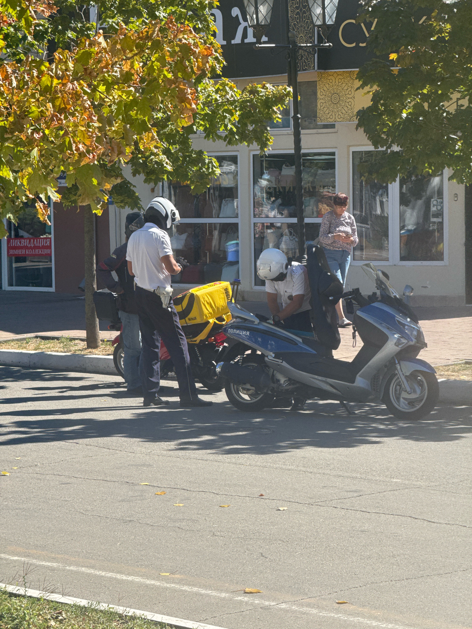 In Anapa, Russia, on a sunny day, two individuals wearing white helmets and dark trousers stand near a blue and silver scooter with "ПОЛИЦИЯ" (Police) visible on its side. One individual, in a white short-sleeved shirt, is interacting with the scooter. A second person, in a dark jacket and blue jeans, stands behind them, also wearing a white helmet. Adjacent to them is a red scooter with a large yellow delivery bag attached to its rear. In the background, a woman in a patterned shirt and blue jeans stands near a building with large windows, holding a mobile phone. Trees with yellowing leaves cast shadows on the asphalt road, which has some scattered fallen leaves. A sign in Russian with "ЛИКВИДАЦИЯ ЗИМНЕЙ КОЛЛЕКЦИИ" (Liquidation of the winter collection) is visible on a building to the left.