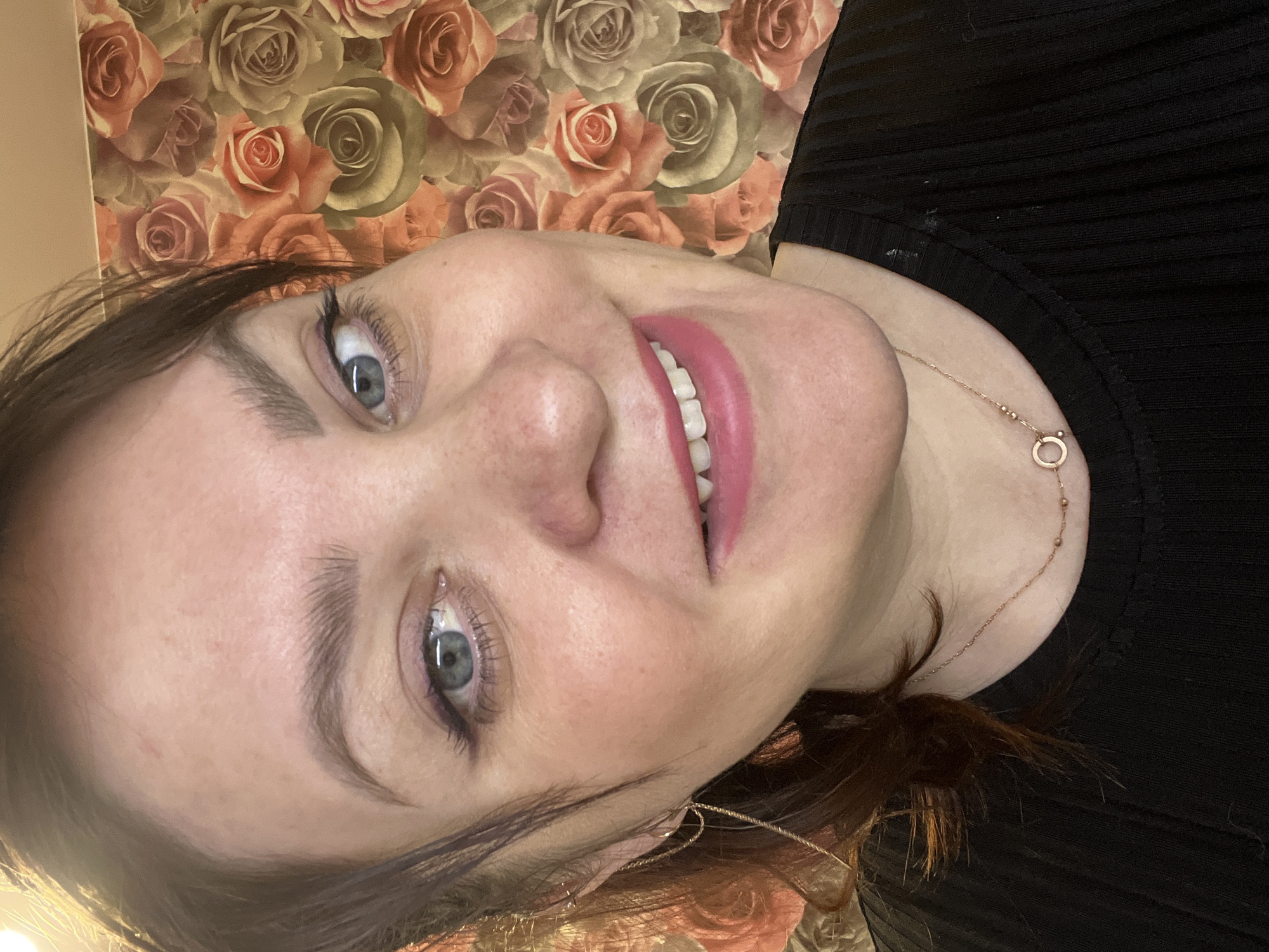 A close-up portrait shows a woman's face, rotated 90 degrees counter-clockwise. She has dark brown hair, blue eyes accentuated with mascara, and well-defined eyebrows. Her skin is fair, with some freckles visible on her nose. She is captured mid-smile, revealing her upper teeth, and wears bright pink lipstick, conveying a cheerful and warm expression.
She is dressed in a black ribbed top, partially visible around her neck and shoulder. Accessorizing her look is a delicate gold-colored chain necklace featuring a small circular pendant, and a thin, gold-colored earring on her visible left ear. The background consists of a patterned wallpaper adorned with numerous roses in shades of peach, orange, and muted green or beige, indicating an indoor setting. The lighting is bright and even, illuminating her features. No text is visible in the image. The scene takes place in Noviy, Russia.