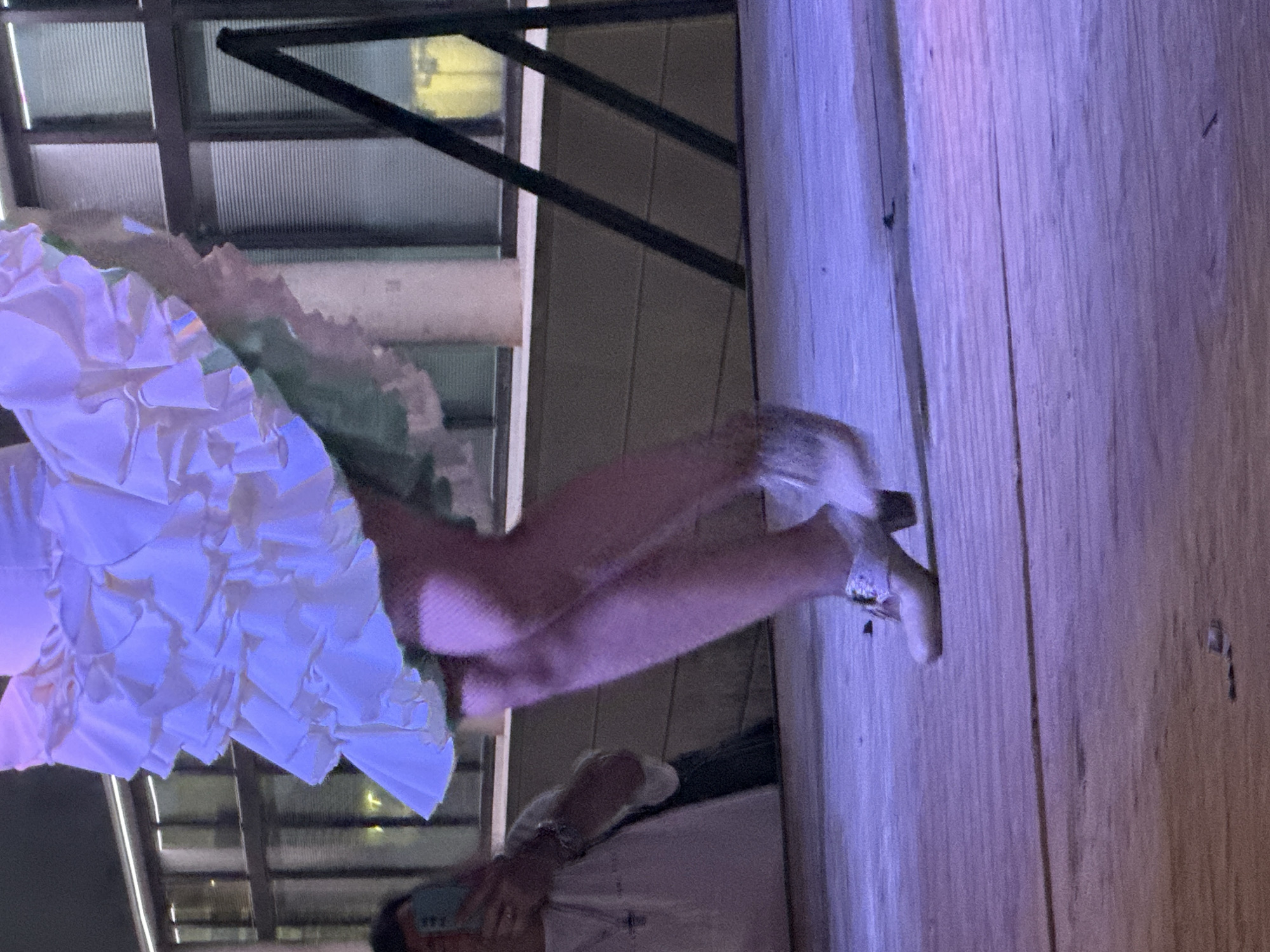 The image shows a close-up of a person's legs and feet on a wooden stage. The person is wearing a ruffled white skirt over a green underskirt and fishnet stockings. Their feet are clad in light-colored heeled shoes adorned with decorative embellishments. In the background, a person is visible in the lower left corner, holding a smartphone. The background also features a building with large windows and some dark, angled structures. The scene is illuminated with purple and blue lighting. The photograph is believed to have been taken in San José Del Valle, Spain, possibly during a performance or cultural event.