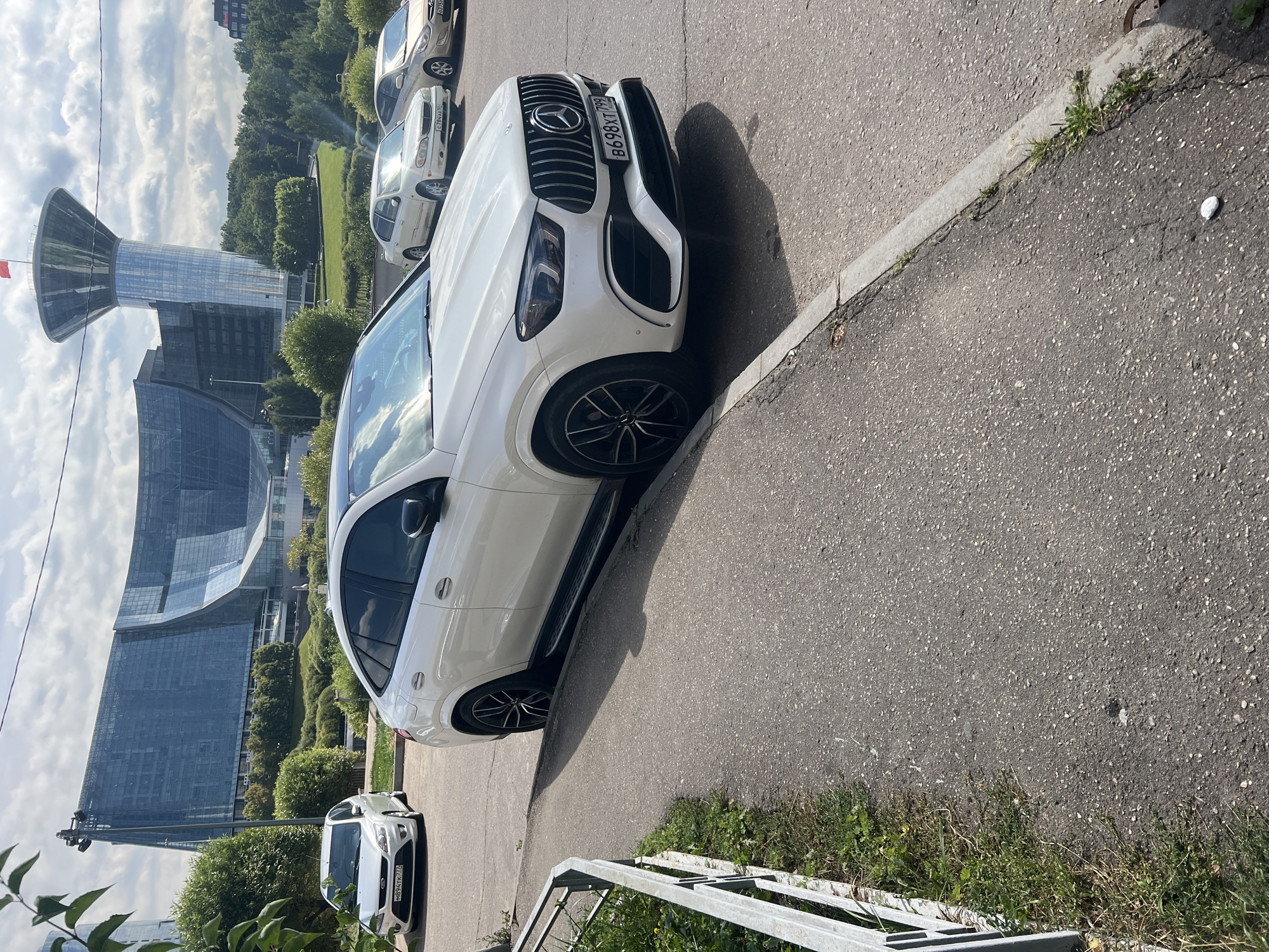 In Khimki, Russia, a white Mercedes-Benz GLE coupe is parked along a paved street. The car has a license plate reading "B698XT 799". In the background, a modern, large, glass-fronted building with an undulating facade is visible, alongside a tall, glass tower topped with a red flag. Trees and green spaces surround the buildings. To the left of the car, a metal fence with a white railing encloses a patch of grass. Other vehicles, including a silver Ford Focus and two sedans, are parked further down the street. The sky is partly cloudy.