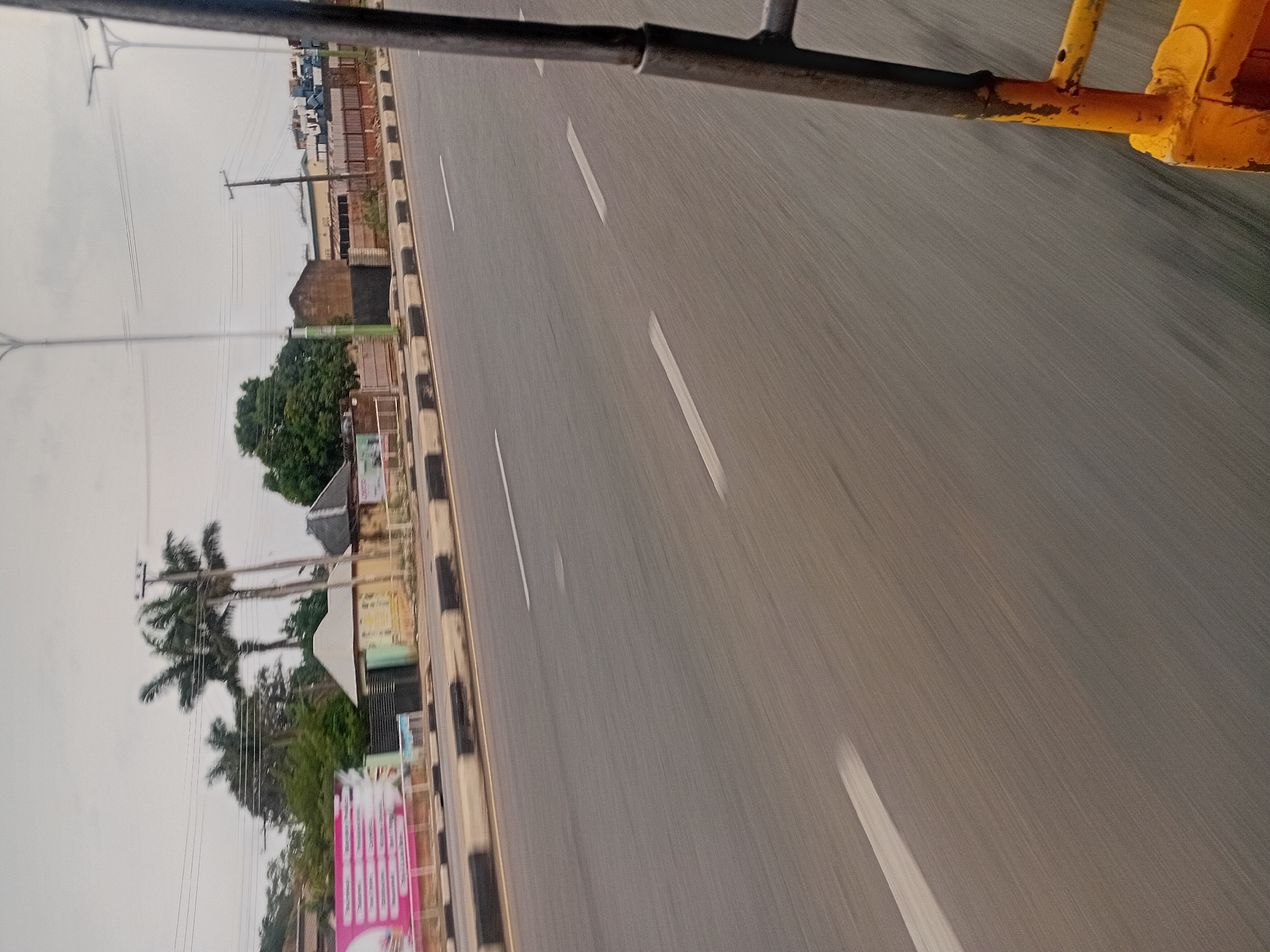 The image shows an asphalt road with white lane markings, possibly in Jos, Nigeria. The road is viewed from a moving vehicle, evident by the motion blur. The roadside features a black and white curb, utility poles, and trees. A partial view of the vehicle's frame, painted black and yellow, is visible in the top portion of the image. Buildings and commercial signs are partially visible along the roadside. The sky is overcast.