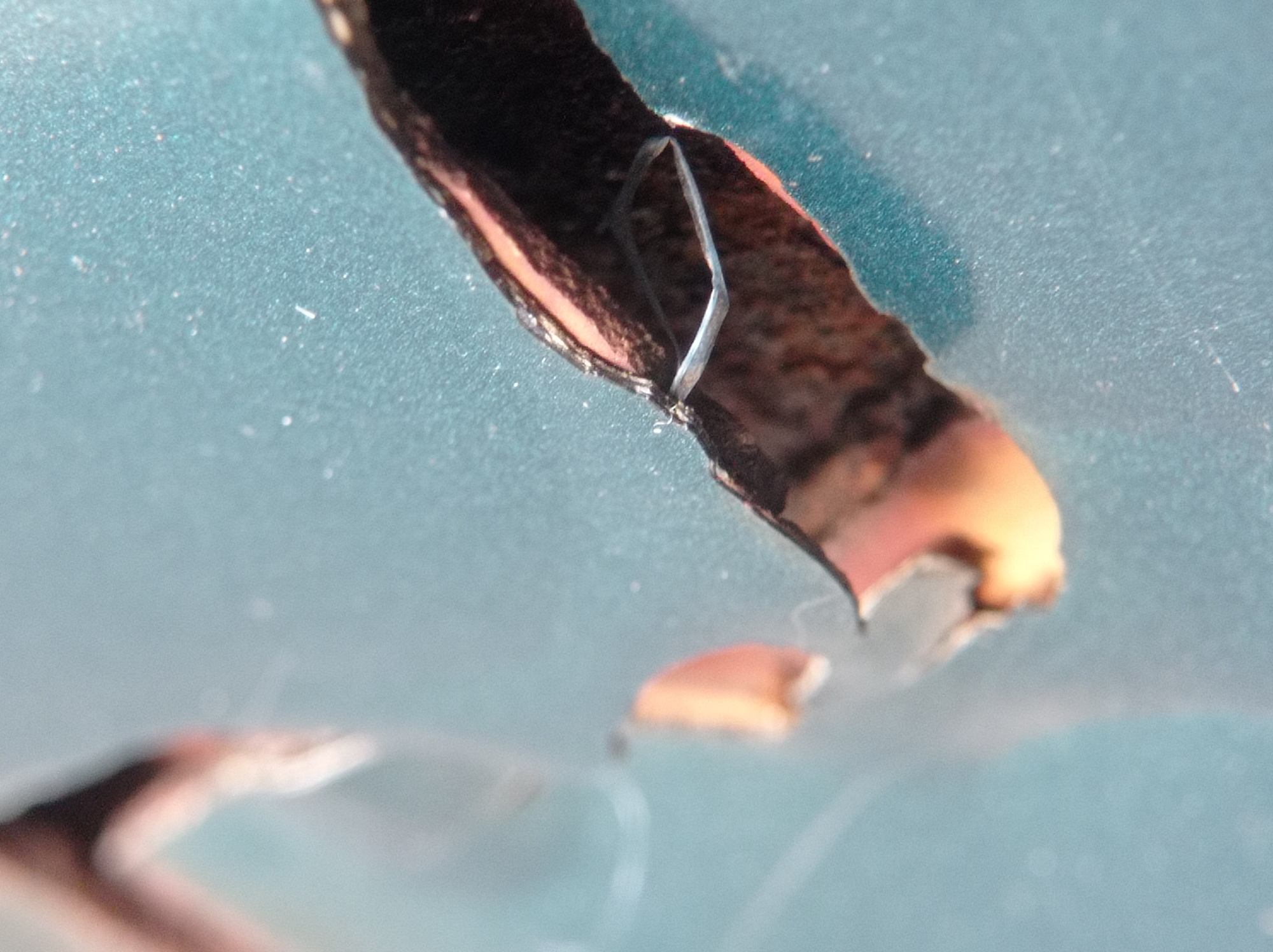 The image shows a close-up of a chipped and peeling surface, possibly paint or a coating, on an object with a teal background. A thin, dark, broken piece of material is detached and curled inward, revealing a light-colored interior. The chipped area has an orange-brown hue beneath the dark outer layer. Small, white particles are scattered on the teal surface. The background is softly blurred, suggesting a shallow depth of field or a textured, matte finish. There are no identifiable landmarks or contextual elements that would place this image in Mulyoagung, Indonesia, or any specific urban environment. The scene is devoid of people or any indication of human activity.
