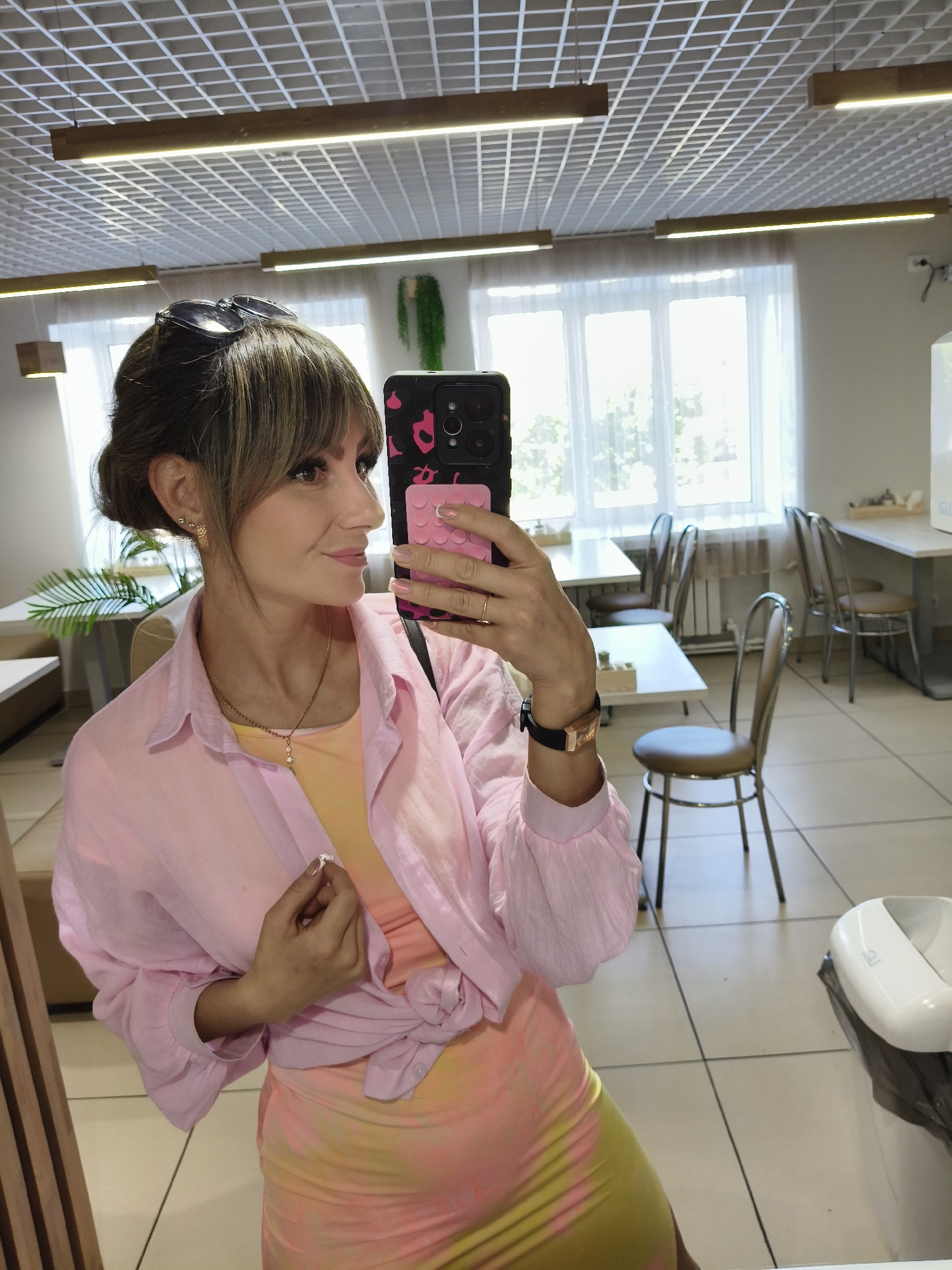 A woman is taking a selfie in what appears to be a café or restaurant in Chaikovsky, Russia. She is wearing a pink shirt tied at the waist over a yellow and pink tie-dye dress. Sunglasses are on her head, and she is wearing a necklace and a watch. Her phone case is black with pink leopard print and a pink square attachment. The background shows tables and chairs, suggesting a dining area. Windows provide natural light.