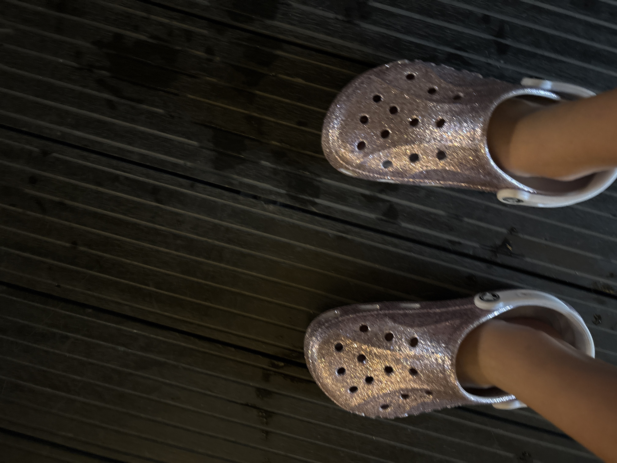 A low-angle, full shot shows a pair of glittery pink Crocs on a person's feet, standing on a dark wooden deck. The deck planks run diagonally across the frame from top left to bottom right. The Crocs have the characteristic holes on the toe box. The person's legs and feet are visible, clad in the Crocs. The scene appears to be outdoors, possibly on a beach or resort, given the proximity to a wooden deck. The lighting suggests it might be evening or a shaded area. The location is identified as Vagator, India.