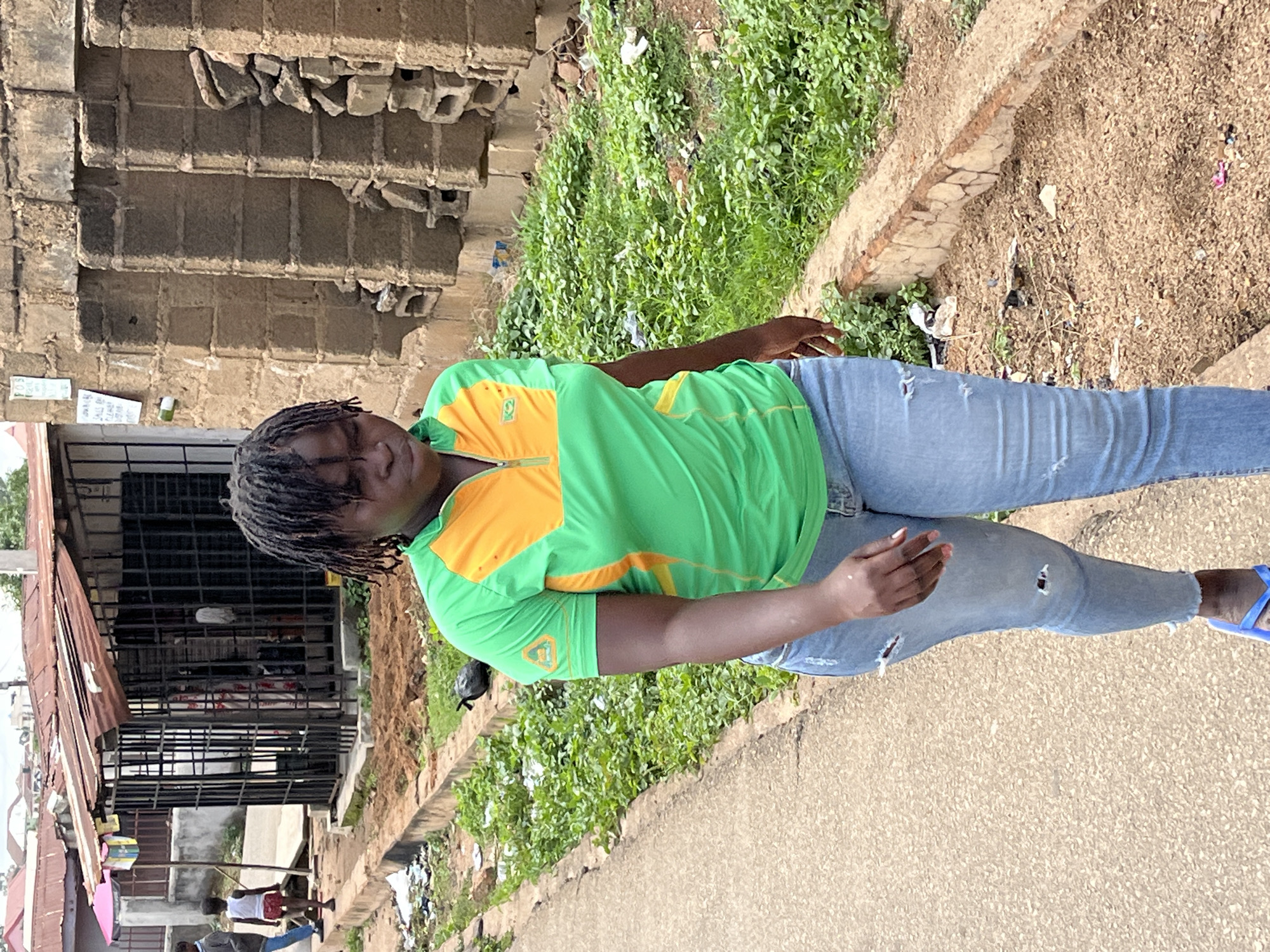 This full-body portrait, taken during daylight hours with bright sunshine, features a person standing outdoors in what appears to be an urban or semi-urban environment in Jos, Nigeria.
The main subject is an individual with dark skin and dreadlocks, wearing a striking green and orange short-sleeved shirt with a zippered collar, light blue ripped jeans, and blue flip-flops. Their left hand has some white markings, possibly paint or powder. They look towards the camera with a relaxed and neutral expression, tilting their head slightly.
The scene includes a paved road or pathway in the foreground, with a concrete curb separating it from a rougher, unpaved area to the left. This unpaved section is covered with abundant green vegetation and sandy patches. In the background, there are various structures. To the immediate left of the subject, a building with exposed and broken blockwork is visible. Further back and to the left, a structure with a corrugated metal roof and prominent metal grates or bars suggests a shop or stall. Another person, possibly a child in a white top, can be seen in the distance near this structure.
Notable details include the bright, clear lighting indicating a sunny day. A small piece of paper affixed to a wall in the background clearly shows the text "JUST BE YOU." The overall setting combines elements of construction, natural overgrowth, and typical street infrastructure, consistent with a dynamic urban landscape.