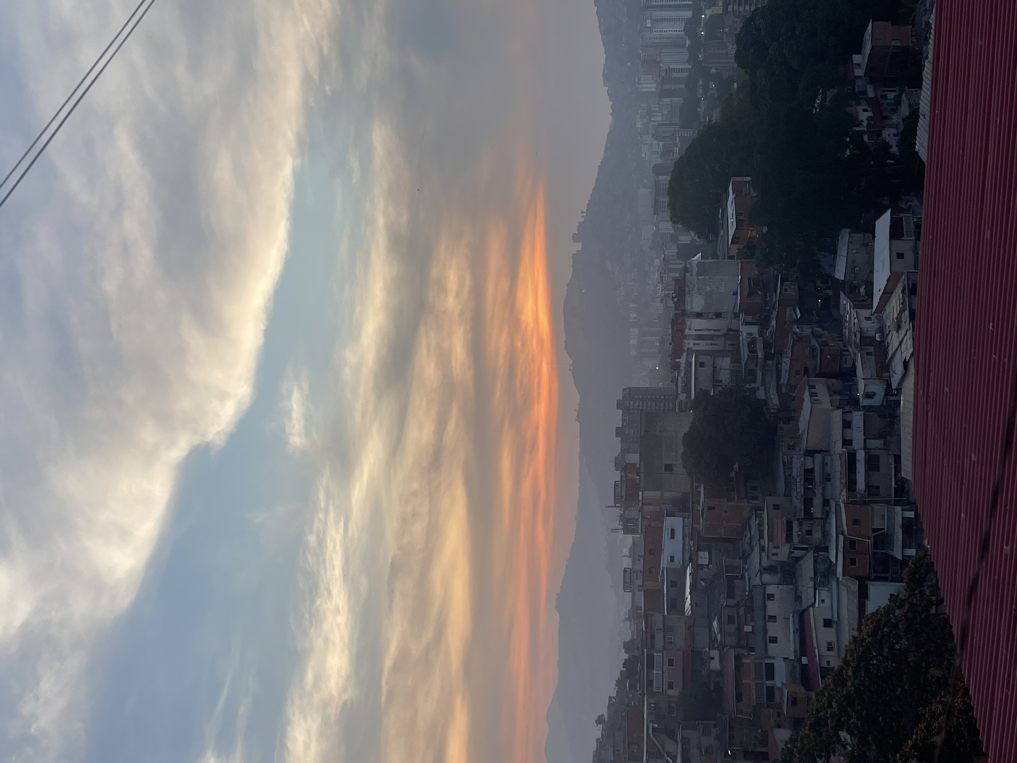 The image depicts an overview of a densely populated area, likely a favela, in Caracas, Venezuela, taken at either sunrise or sunset.
**Environment**: The sky shows a gradient of light blue to a band of orange and yellow hues, suggesting either dawn or dusk. The air is hazy, obscuring the distant mountains.
**Subjects and Objects**:
* **Buildings**: The scene is dominated by tightly packed buildings of varying heights and colors. The structures appear to be informal housing.
* **Vegetation**: Patches of green trees intersperse among the buildings.
* **Sky**: The sky contains streaks of light blue and grey-white clouds.
* **Foreground**: In the upper-left corner, power lines are present. On the right side, a red corrugated iron roof is partially visible.
**Location**: Based on the dense urban development nestled against hilly terrain, it is characteristic of the informal settlements found in Caracas, Venezuela.
**People/Actions**: No people are directly visible in the scene, suggesting a view from a distance or a time when outdoor activity is low.