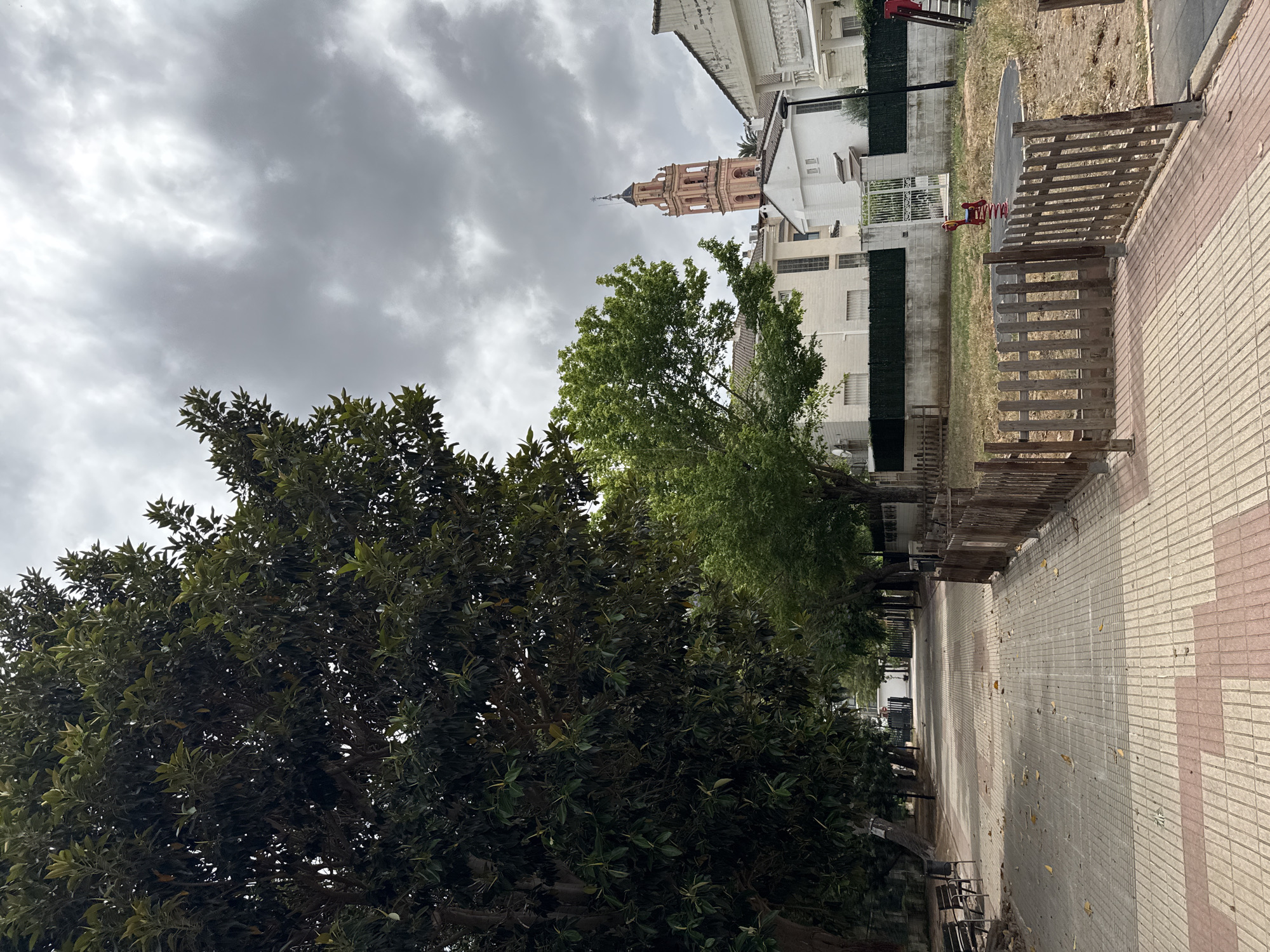 A tree-lined pathway stretches into the distance under a cloudy sky in Benirredrà, Spain. On the left, a large, mature tree with dark green leaves dominates the foreground, its branches reaching towards the overcast sky. To the right of the pathway, a small, fenced-off playground area contains a red spring rider. Beyond the playground, houses with white facades and tiled roofs are visible. In the background, a light pink bell tower with arched openings and a dark steeple rises above the buildings, indicating a church or similar structure. The pathway is paved with rectangular tiles in a checkerboard pattern of beige and red. Benches are placed along the left side of the path. The overall scene depicts a public space with residential elements and a touch of recreational area.