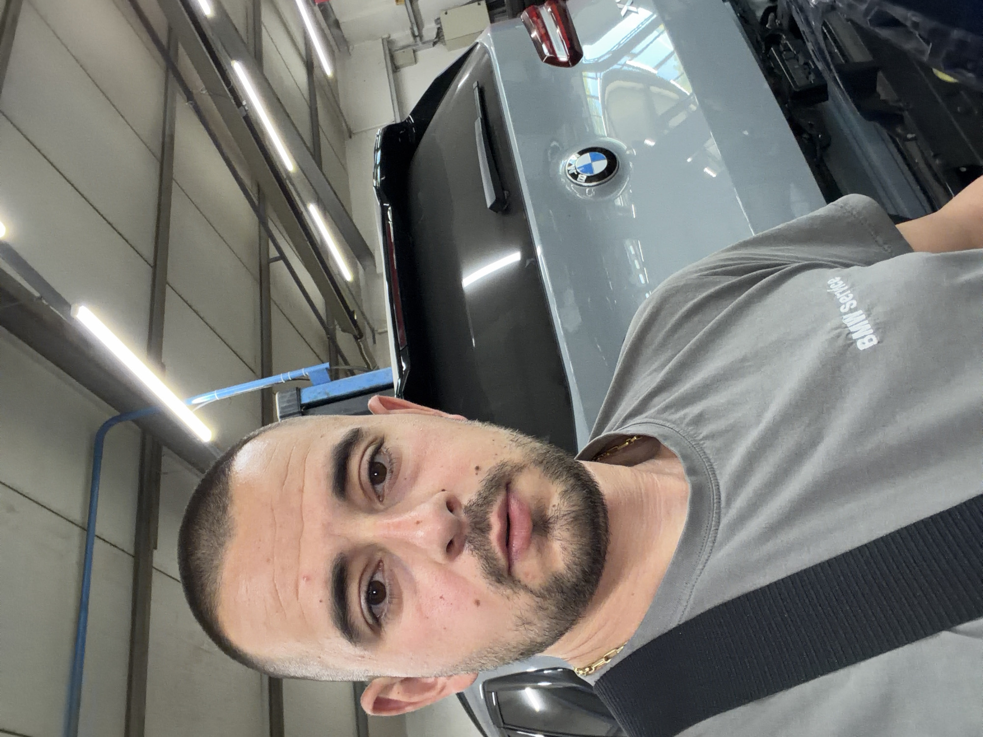 The image shows a man standing in front of a grey BMW SUV in what appears to be a car service center. He is wearing a grey t-shirt with "BMW Service" printed on it and a black strap across his chest. The man has short hair, a beard, and is looking at the camera. The background shows fluorescent lights on the ceiling and other elements of a workshop. The car has a BMW logo on the back. The location is identified as Burgas, Bulgaria.