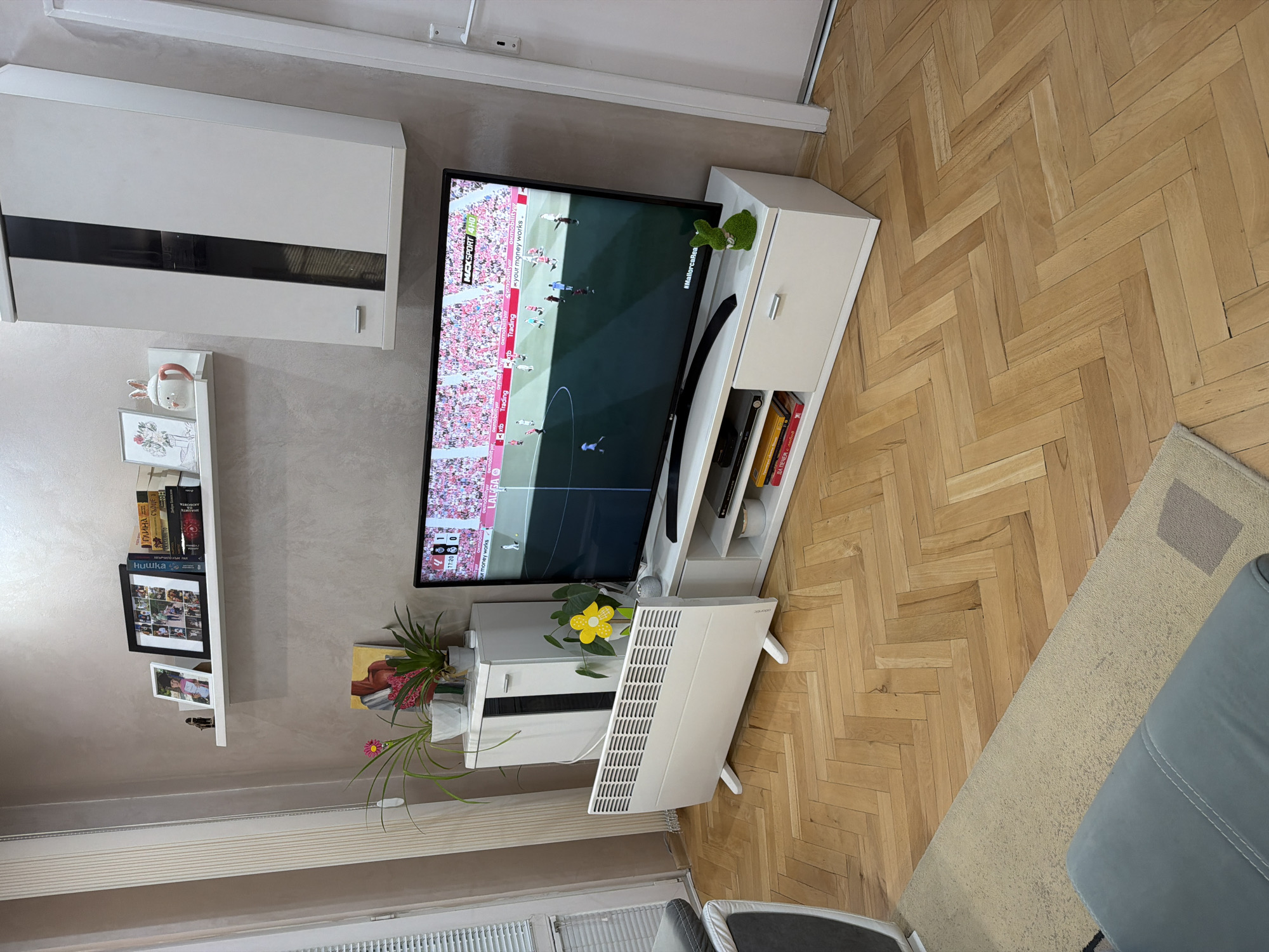 Here is a concise description of the image:
The image shows an interior view of a room with herringbone wood flooring. A television displays a soccer match. Adjacent to the TV is a white shelving unit. There's a white cabinet mounted on the wall with a bookshelf and decor below. A portable heater stands near a window with closed blinds.
I'm unable to determine any details related to the location (city of Plovdiv, Bulgaria) as there is no forensic data in the image.