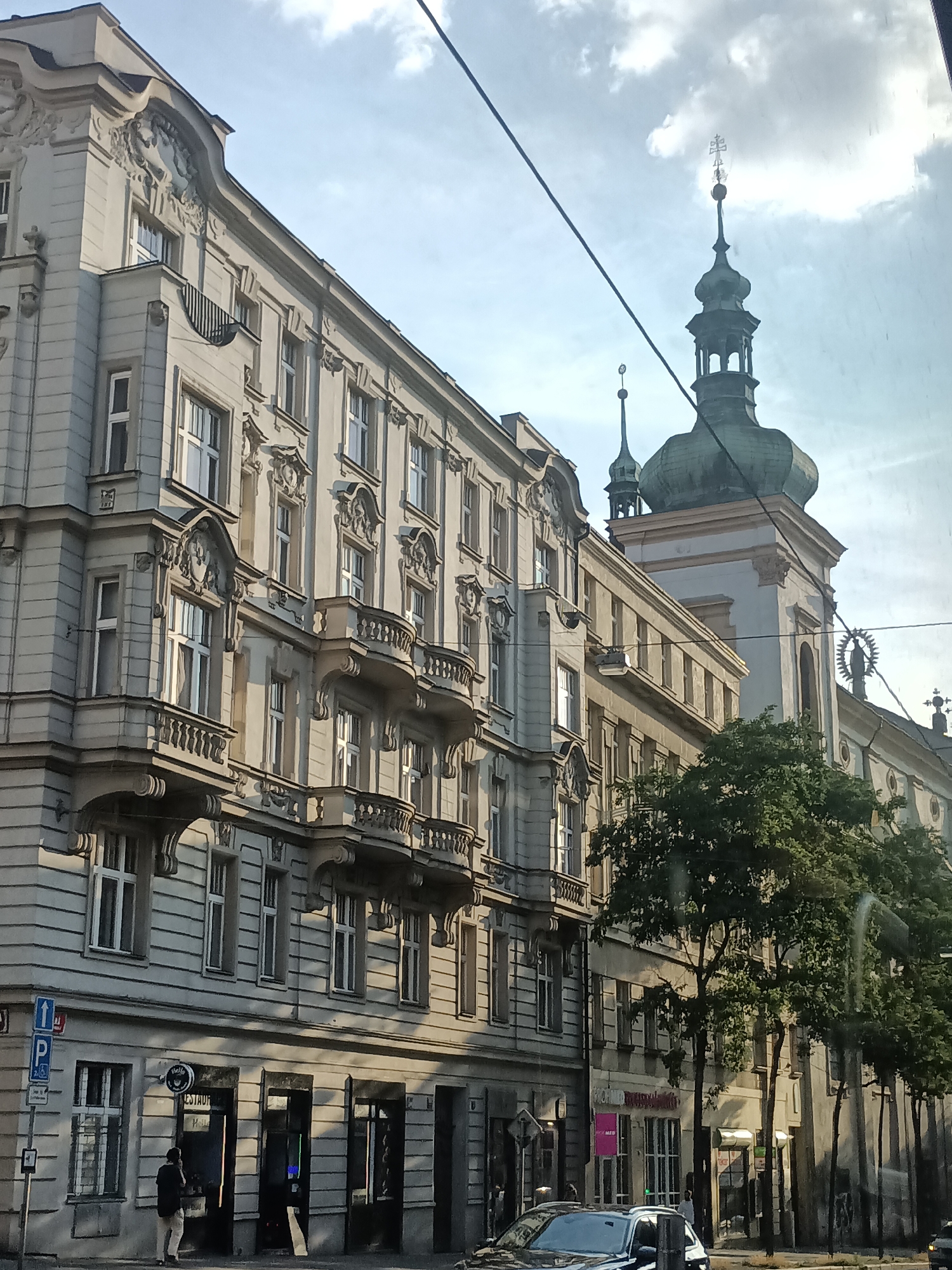 The image shows a street scene in Prague, Czechia. On the left is a multi-story building with ornate architectural details, including balconies with decorative railings. The building has many windows and a pale facade. To the right of this building is a church with a green dome and a spire, topped with a cross. The church has a tall, square tower and appears to be connected to other buildings. Trees with green foliage line the street in front of the church and other buildings further down. The sky is partly cloudy. In the foreground, there is a black car and a person standing on the sidewalk, looking at their phone. There are also street signs and what appears to be the entrance to a restaurant. Power lines are visible stretching across the upper part of the image. The lighting suggests it is daytime, likely late afternoon given the shadows.