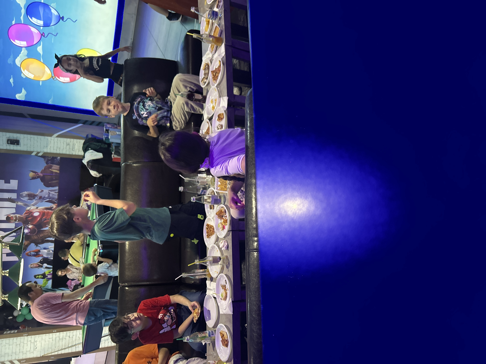 A group of people, mainly children, are gathered around a long table in what appears to be an entertainment venue in Burgas, Bulgaria. The table is set with plates, some containing food, and bottles with straws. People are eating and talking. In the background is a large screen displaying images and balloons. The location seems to be indoors with artificial lighting. The venue displays various decorations including video game and superhero themes, indicative of a child-friendly or family-oriented business.