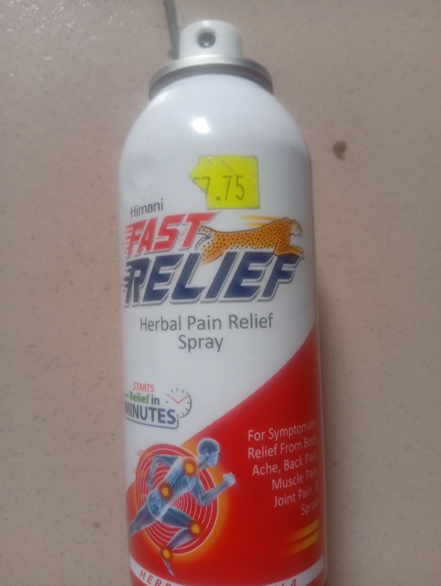 The image is a close-up shot of a can of Himani Fast Relief Herbal Pain Relief Spray. The can is primarily white with red and blue lettering. A yellow sticker with the number "7.75" is affixed to the upper portion of the can. The background appears to be a speckled, beige surface. The can is positioned vertically, and the lighting suggests an indoor setting. There is no indication of the city of Ashata, Nigeria, in the image or its context.