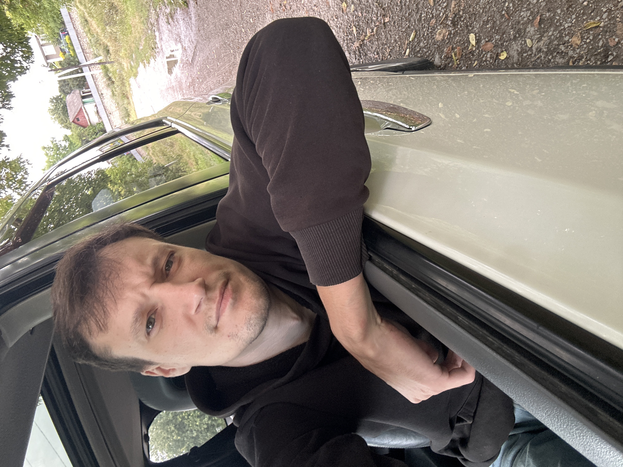 A Caucasian male is captured in a selfie from inside a car, with his right arm resting on the open car door. He is wearing a dark hooded sweatshirt and blue jeans. The background reveals a rural setting with a dirt road, some trees, and a few houses. The location is identified as Makiv, Ukraine. The scene appears to be outdoors, with overcast lighting.