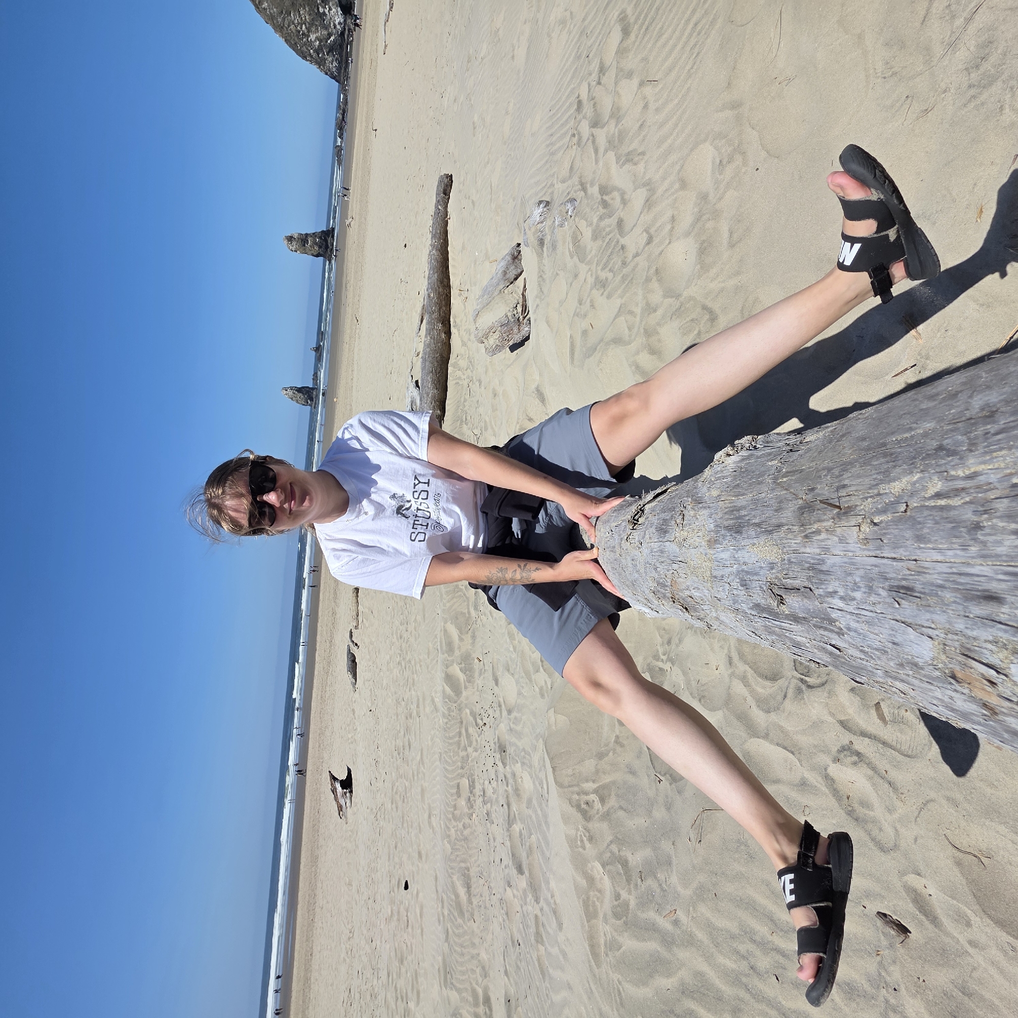 A young woman with blonde hair, wearing sunglasses, a white t-shirt with a "STUSSY" logo, and gray shorts, sits on a log on a sandy beach. Her legs are spread wide, and her hands rest on the log. In the background, the ocean meets the shore with gentle waves, and several rock formations rise from the water. The sky is clear and blue. The image appears to be taken at Cannon Beach, Oregon, United States of America.