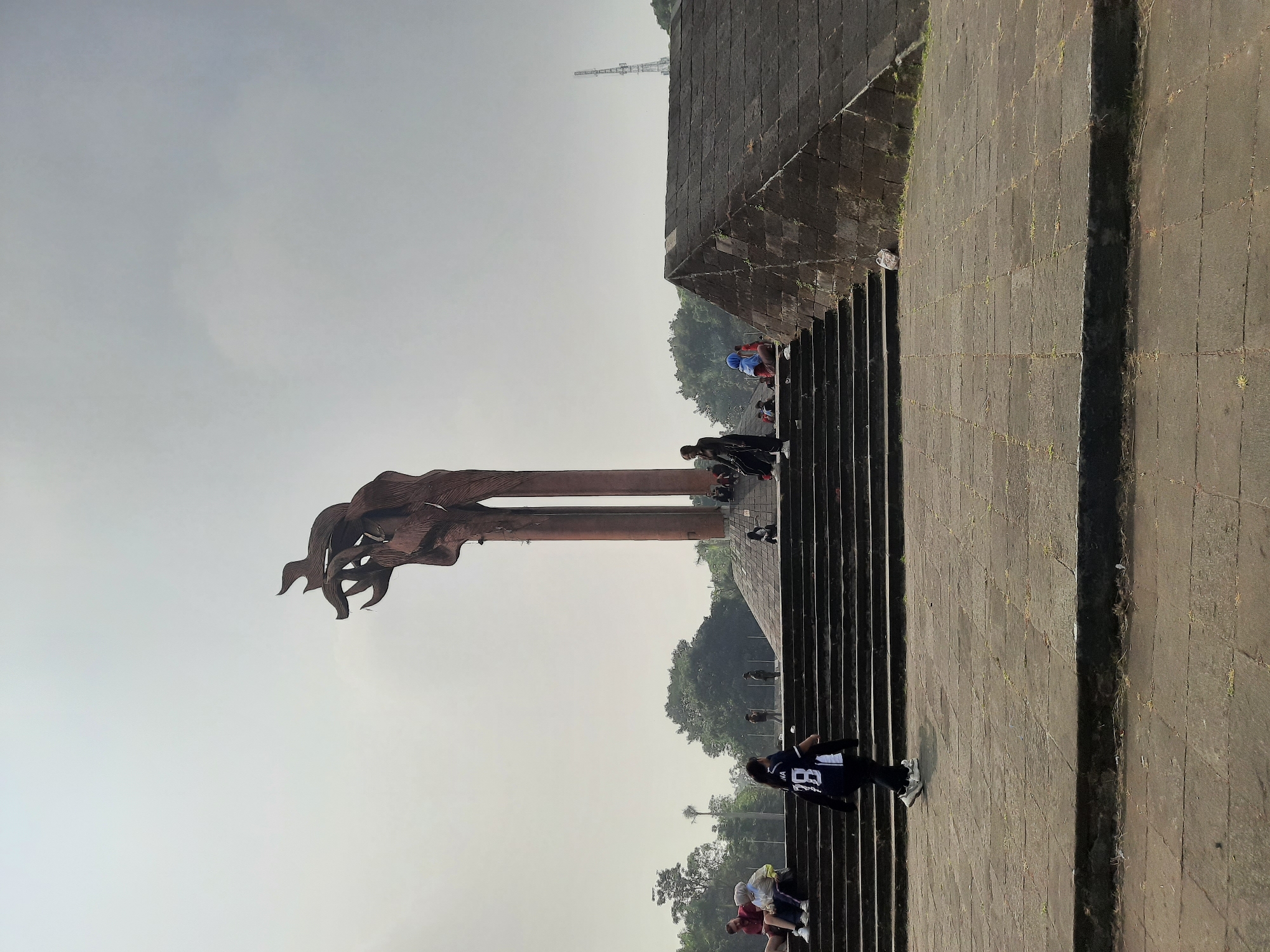 A wide-angle shot captures a monument in Bandung City, Indonesia, under a hazy sky. The monument, which appears to be made of metal, features three tall, upward-curving structures that resemble flames or abstract figures. The base of the monument is surrounded by a large, tiered stone plaza.
In the foreground, a wide stone staircase leads up towards the monument. Several individuals are present on the stairs and plaza. On the lower steps, a young woman wearing a dark jersey with the number "18" is walking away from the camera. Further up the stairs, other people are seated or standing, some in casual conversation. To the right of the stairs, a large, dark stone pyramid-like structure dominates the mid-ground.
The environment is characterized by a park-like setting with lush green trees visible in the background. A tall communication tower is also present on the horizon. The overall scene conveys a sense of public gathering and leisure in an urban park.