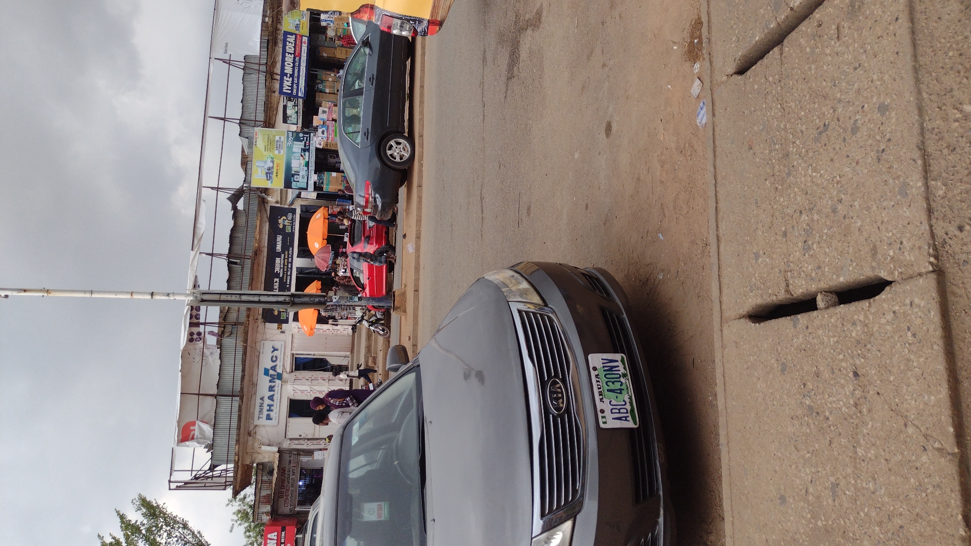 The image depicts a street scene in what appears to be a commercial area, likely in Jos, Nigeria. A gray Kia car with license plate "ABC-430NV" partially occupies the foreground. A dark-colored car is parked further down the street. Pedestrians and possibly vendors are visible near storefronts. Signage for businesses such as "TINNA PHARMACY" and "IYKE-MORE IDEAL" are present. The environment is overcast with a cloudy sky. The street has a concrete curb with drainage openings.