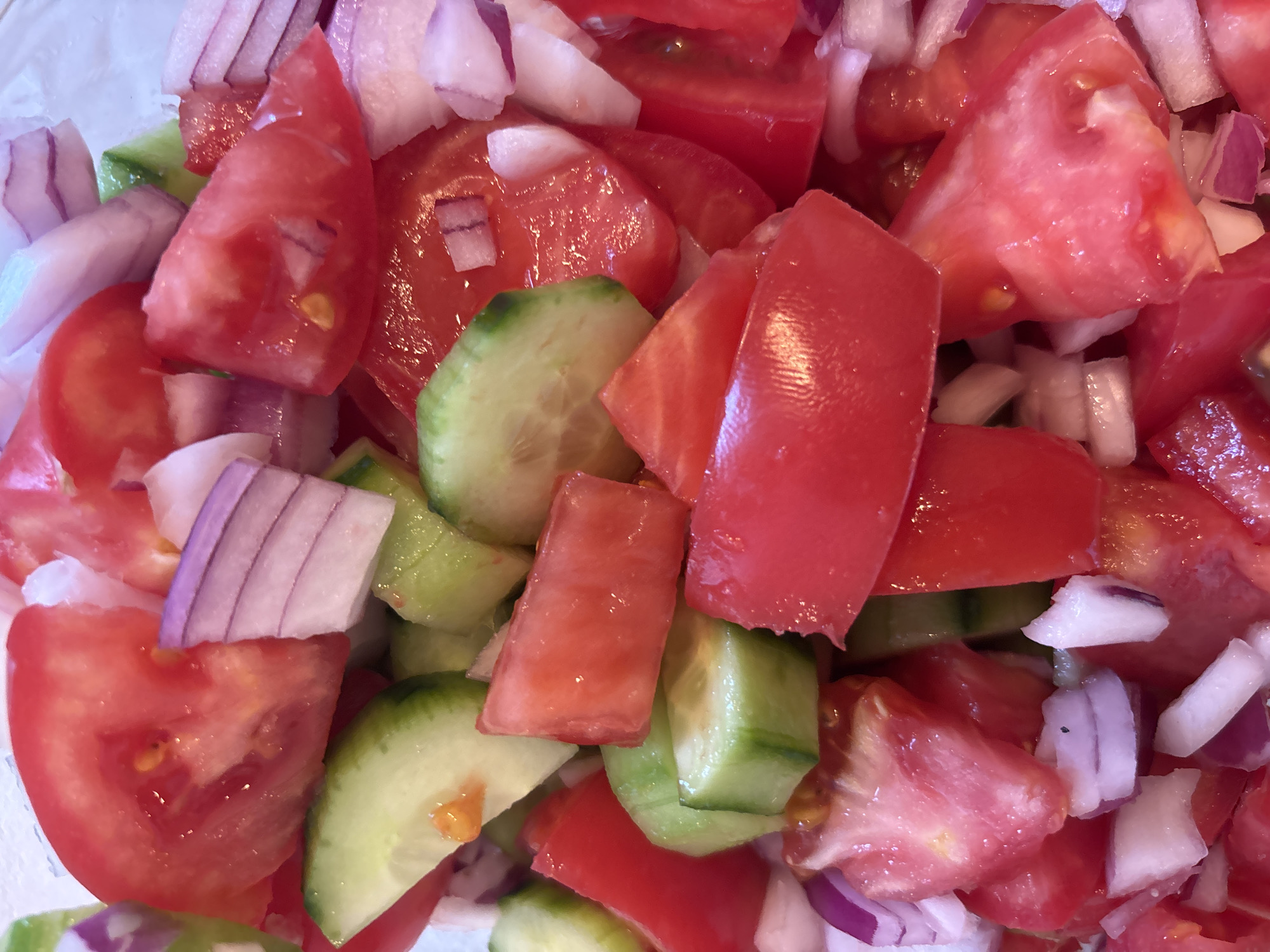 A close-up, overhead shot shows a chopped tomato and cucumber salad with red onion. The salad is in a white bowl. The tomatoes are sliced into wedges, and the cucumbers are sliced into rounds. The red onion is chopped into small pieces. The salad appears to be fresh and ripe. The lighting is bright and natural, suggesting the salad is outdoors or near a window. There are no people or animals visible in the image. The background is blurred, focusing the viewer's attention on the salad.