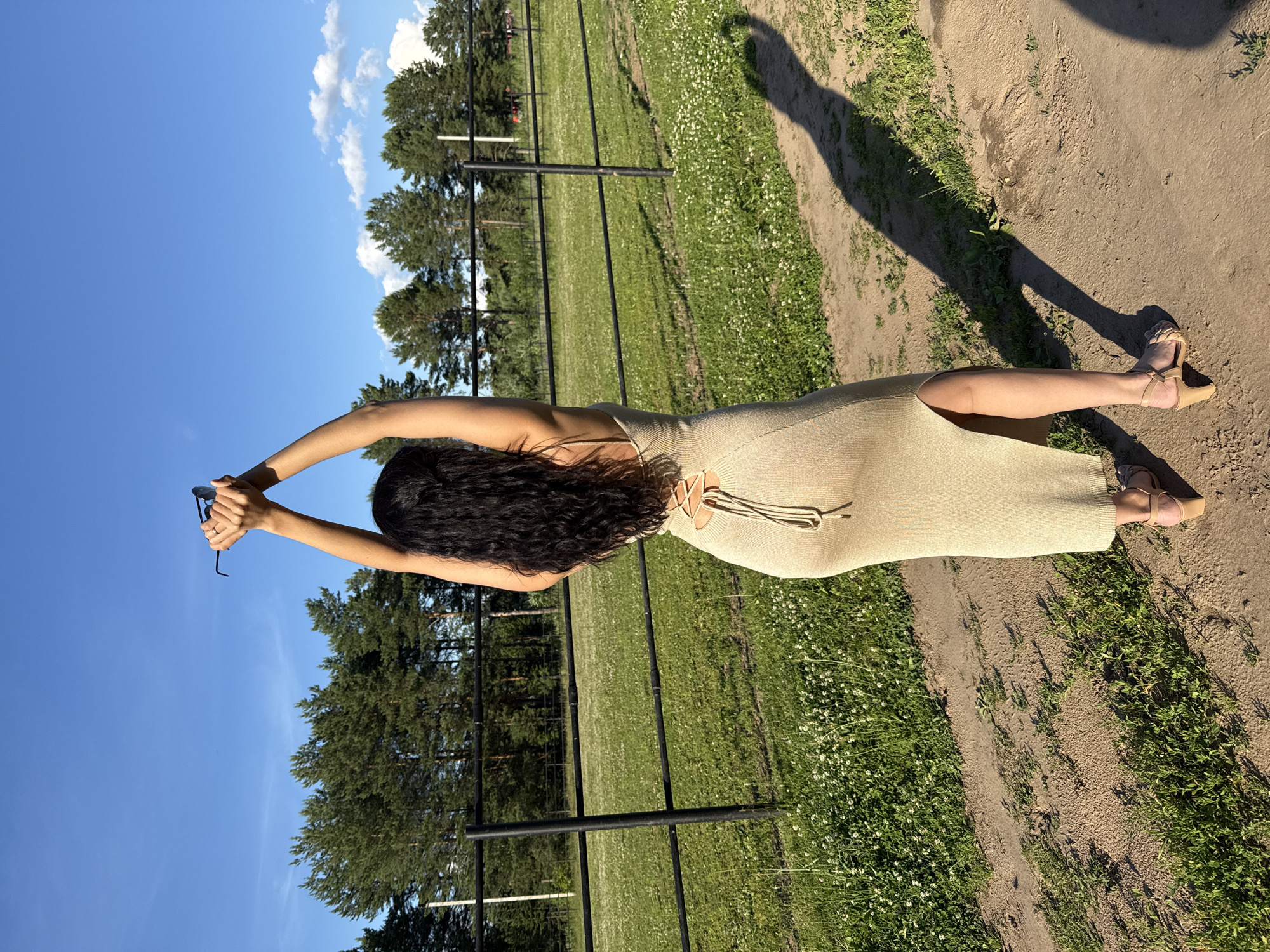 In Chaikovsky, Russia, a woman with long dark hair stands on a dirt path facing away from the camera. She is wearing a light-colored, form-fitting, floor-length dress with a high slit on the side and a lace-up detail on the back. Her arms are raised above her head, and she is holding a pair of sunglasses. She is wearing light-colored sandals with a heel. The background features a black fence, a grassy field with scattered white flowers, and a line of coniferous trees under a bright blue sky with some scattered clouds. The lighting suggests it is daytime, likely late afternoon due to the long shadow cast by the woman.