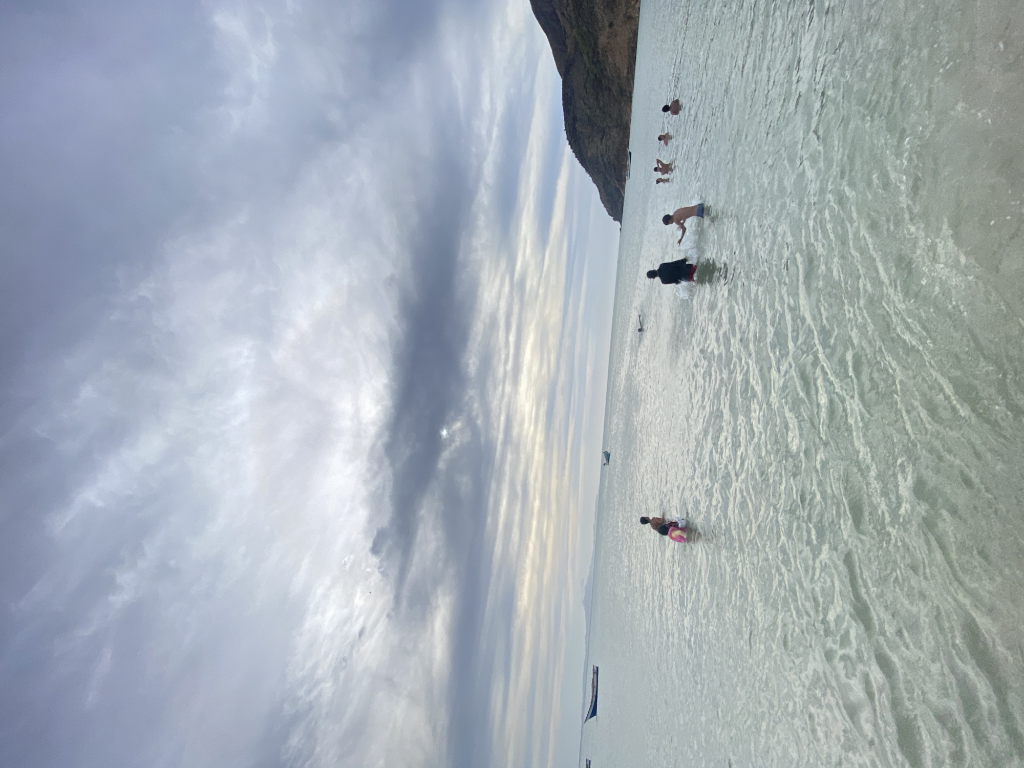 The image shows a coastal scene with several people wading in shallow, clear water. A dark, rocky headland is visible in the background. The sky is filled with streaky, textured clouds. Some boats are in the water.
This is in Juan Griego, Venezuela.