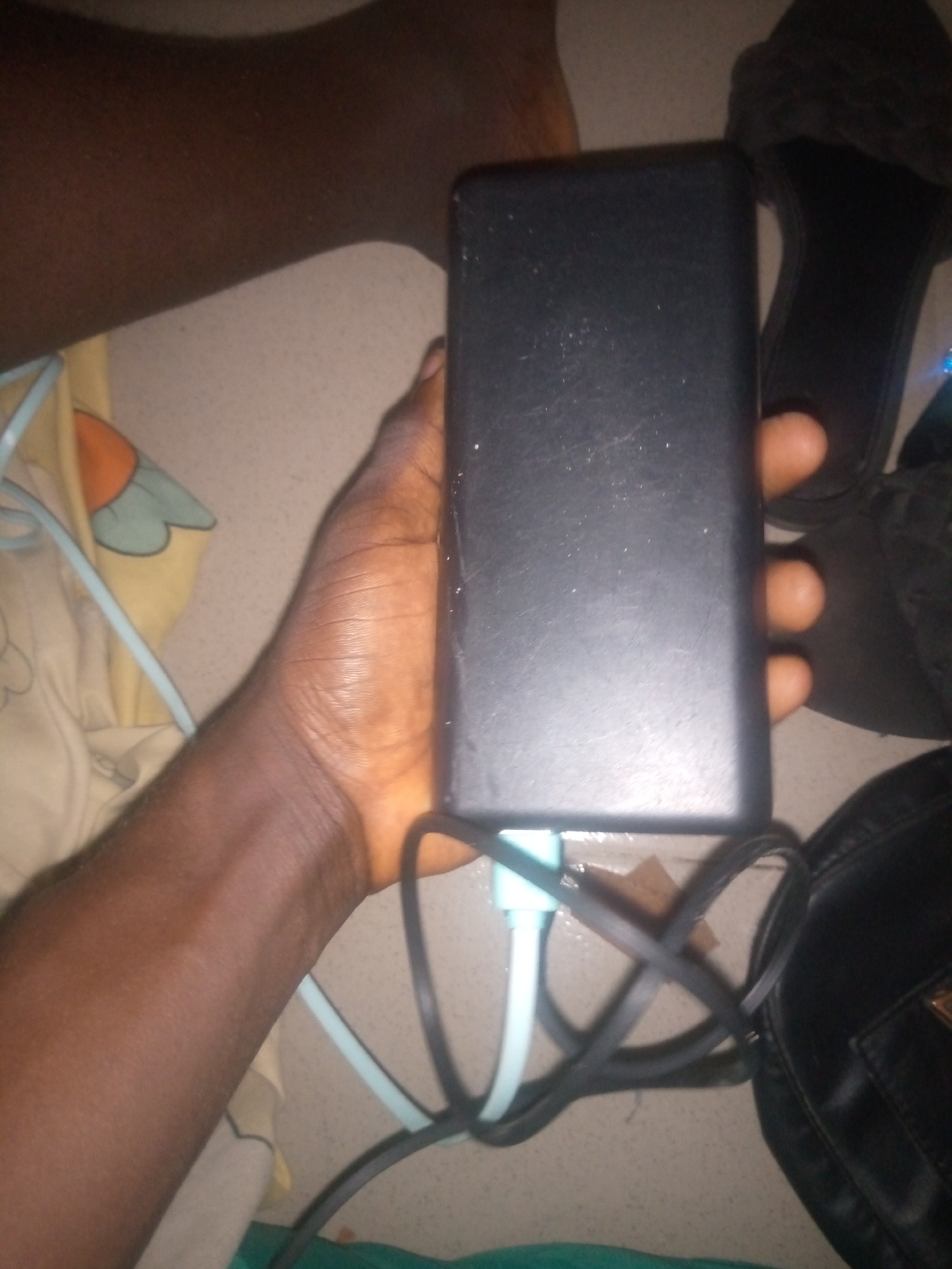 An overhead, low-angle medium shot shows a person's right hand holding a black rectangular power bank connected to a light blue USB cable. The power bank is positioned vertically in the center of the frame, with the cable emerging from its bottom. The person's fingers are visible, wrapped around the power bank and the cable. To the right of the power bank, a black sandal is visible, and further to the right, a portion of a black handbag is present. In the background, a person's left foot and leg are partially visible, along with a light-colored fabric with a yellow and green pattern. The scene appears to be indoors on a tiled floor.