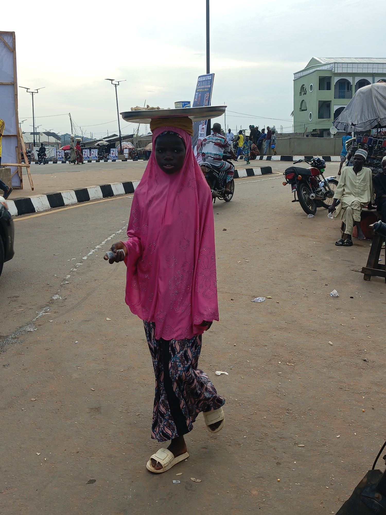 In Akwanga, Nigeria, a young girl wearing a vibrant pink hijab and patterned skirt walks across a dusty, paved road. She carries a large flat tray balanced on her head, holding food items or goods. In her right hand, she holds a small white object, possibly money or an item for sale. The road has a striped curb marking. In the background, other people are visible, some on motorcycles and others standing near market stalls. A light green, multi-story building stands in the distance, and several streetlights are present. The sky is overcast. The scene depicts a busy, everyday street in Akwanga.