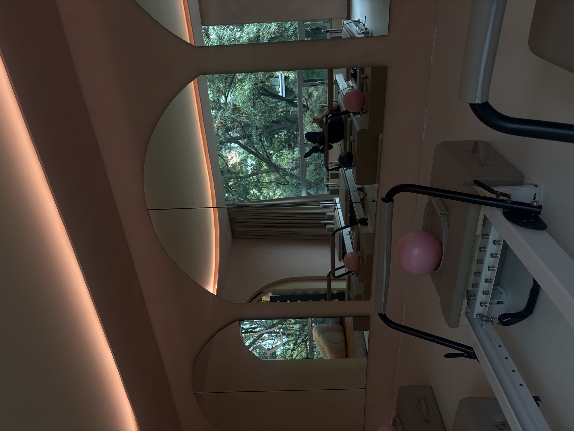 The image shows a room with a pinkish-peach tone and unique architectural design. Multiple arched mirrors reflect outdoor scenery, with trees visible through large windows. These mirrors reflect a person seated in a pilates machine, likely in a studio setting. Additional pilates equipment is present in the foreground, specifically a reformer with a pink exercise ball attached. The exact location within Mexico City is not discernible from the visual information available.