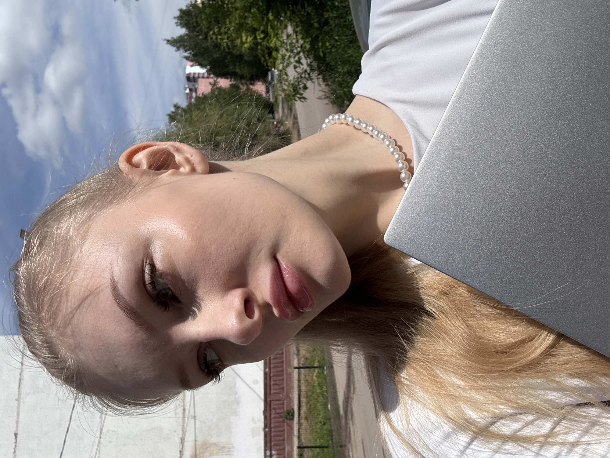 A close-up, rotated outdoor portrait features a young woman with light blonde or light brown hair and fair skin. Her head is tilted, and she looks downwards with a neutral to slightly contemplative expression. Her lips appear glossy, and she has soft eye makeup. She is wearing a white top and a pearl necklace around her neck.
The upper part of a silver or gray laptop is visible in the lower right portion of the frame, partially obscuring her body. Bright sunlight illuminates her face and hair, creating some highlights.
In the background, a clear blue sky with sparse white clouds is visible. Lush green trees and parts of distant buildings, including one with pink and white elements, suggest an urban or suburban outdoor setting, likely in Perm, Russia, during the daytime. The overall impression is a casual, sunlit moment. No visible text is present in the image.