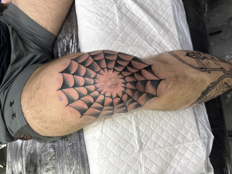A person's leg features a spiderweb tattoo on the knee. The design is black and gray, centered directly on the kneecap. The leg is also adorned with another tattoo higher up. The person is wearing gray shorts with an Under Armour logo, and the leg is resting on a white, padded surface.