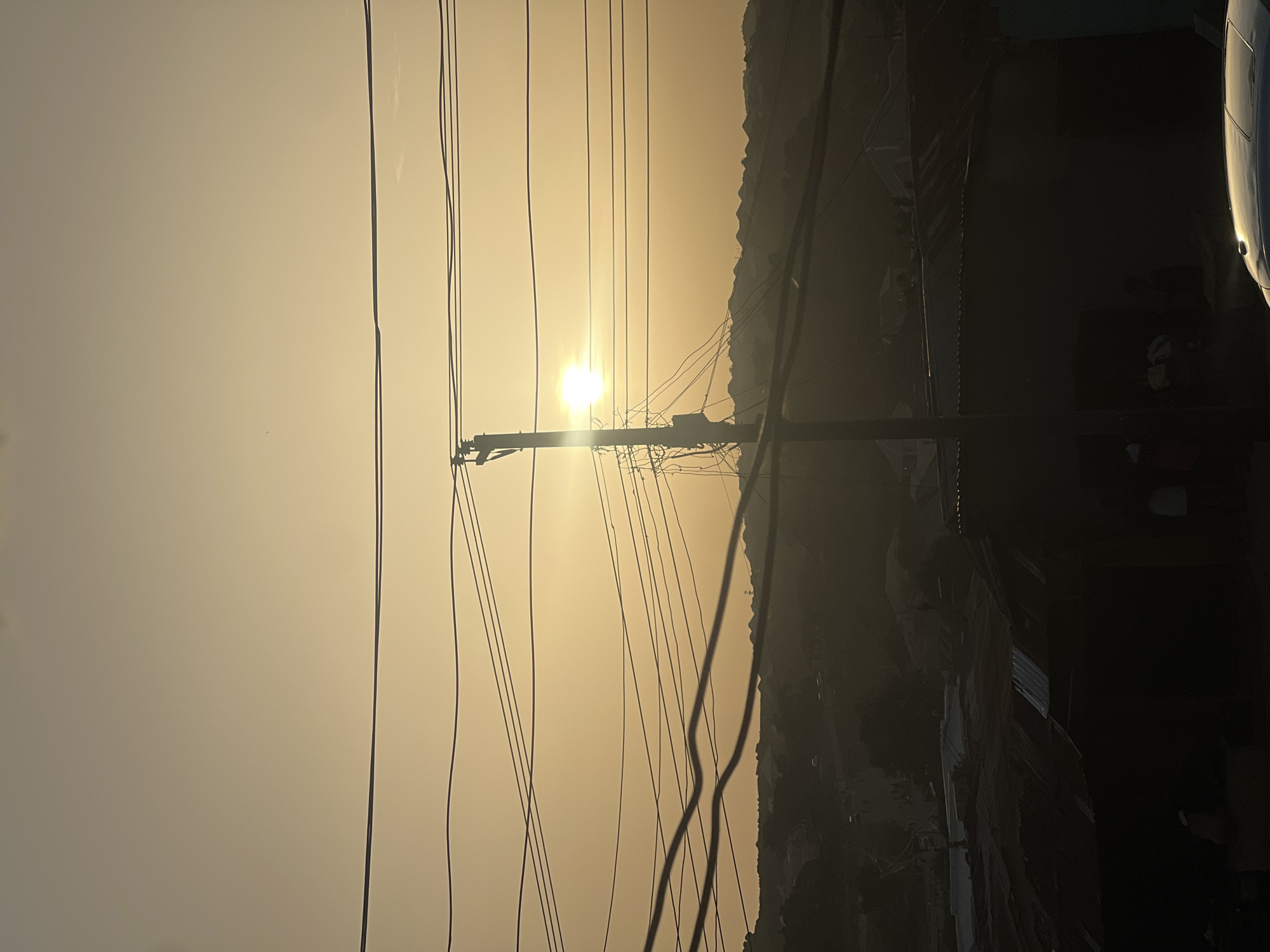 The image depicts a street scene in Jos, Nigeria, at dusk or dawn, with a hazy, golden sky. A utility pole is centrally located, with numerous power lines crisscrossing the frame. Buildings and/or natural rocky outcrops are visible in silhouette along the right side of the image. A portion of a vehicle is visible in the far upper-right corner. No people are immediately visible. The overall atmosphere is obscured by dust, haze, or smoke.