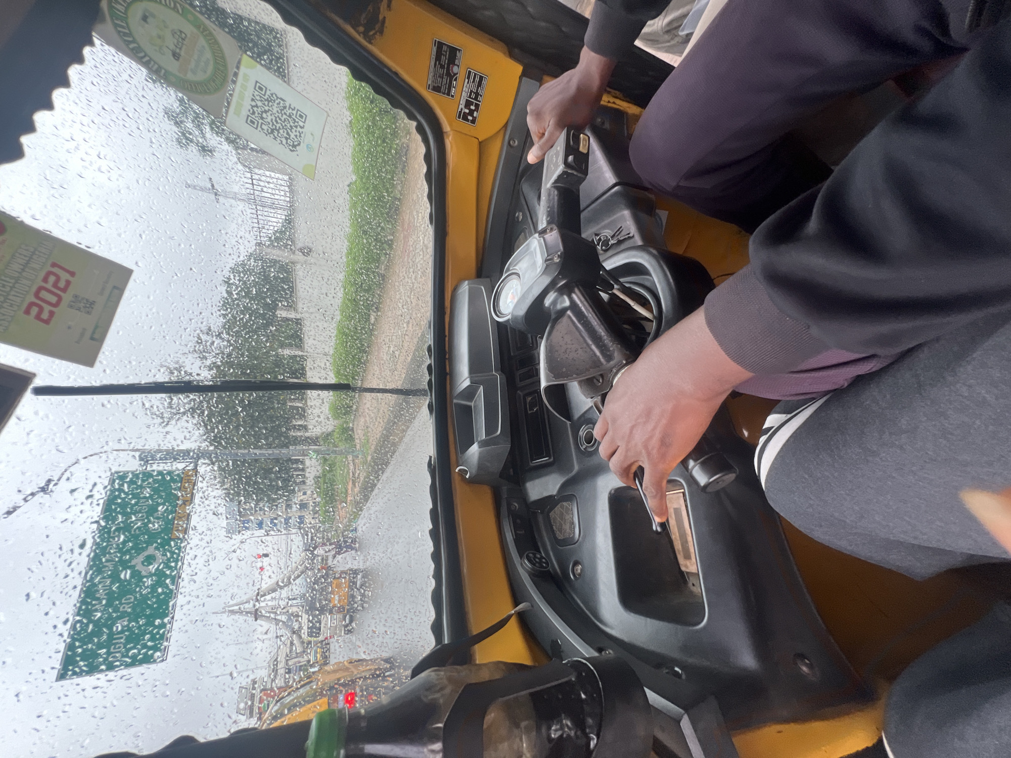 The image shows the interior of a yellow tricycle, commonly known as an "Okada" or "Keke Napep," in Onitsha, Nigeria. The view is from the driver's perspective, looking through the windshield, which is covered in raindrops, indicating a rainy day. The driver's hands are visible on the steering wheel and controls. In the foreground, parts of the driver's and passenger's legs are visible, wearing casual clothing.
Through the windshield, a busy street scene unfolds. Traffic is moderate, with other tricycles and vehicles on the wet road. A prominent green sign is visible on the left, indicating "Main Market" and "Oguya Road" with a roundabout symbol, suggesting the location is near a major market area in Onitsha. Various stickers, including one from "Motorcycle Owners Association of Nigeria 2021" and another with a QR code, are affixed to the interior of the windshield. The surrounding environment shows trees and buildings, typical of an urban setting. The overall impression is of navigating through city traffic in wet weather conditions.