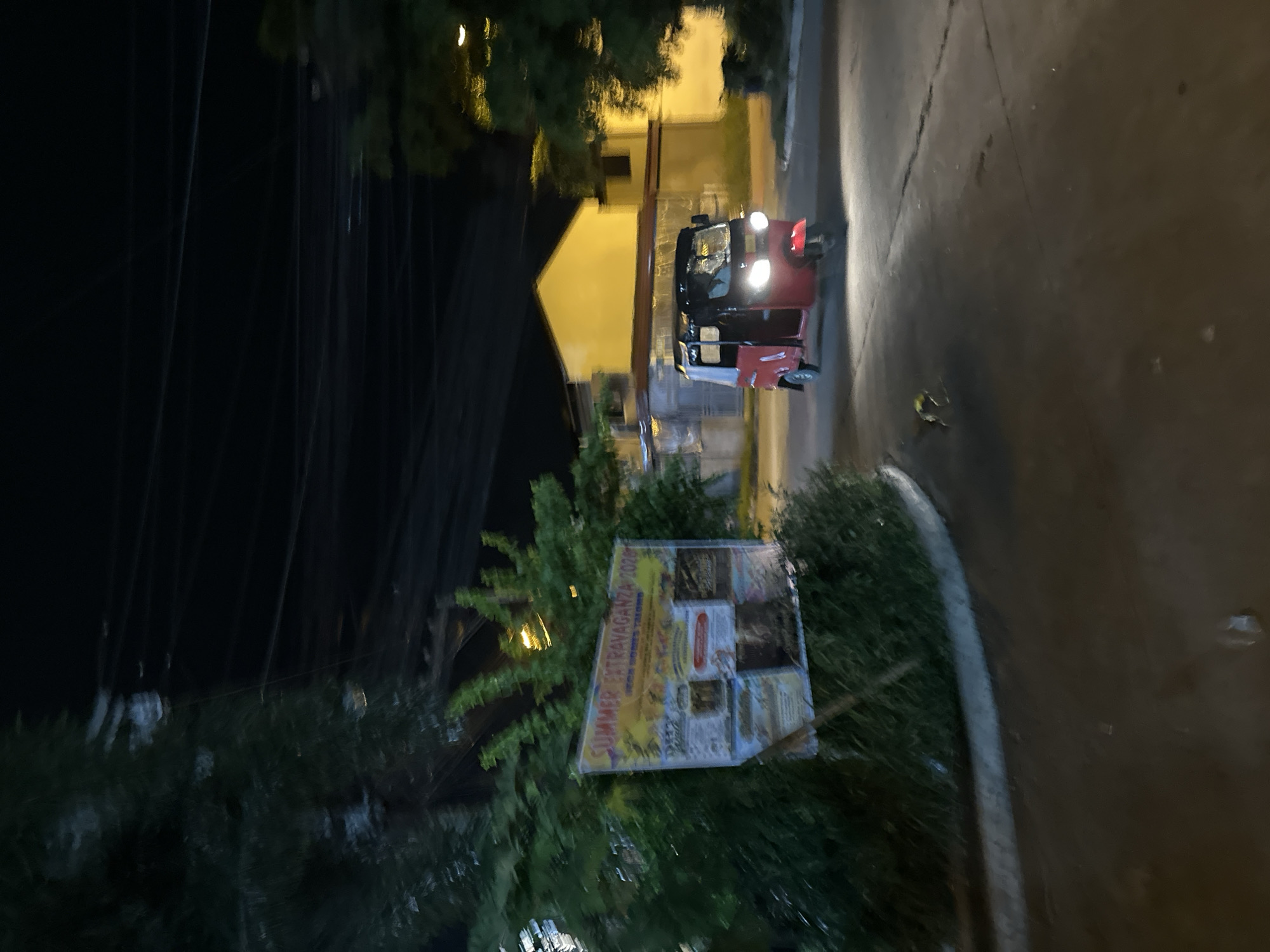 The image depicts a nighttime street scene in Davao City, Philippines, characterized by artificial lighting and local transport. A prominent red and black tricycle, a common auto-rickshaw vehicle in the region, is captured with its headlights brightly illuminated as it moves along a paved road. The road surface appears dark but is partially lit by the tricycle's lights and ambient street illumination.
In the background, buildings with warmly lit, yellow-toned walls are visible, suggesting nearby residences or commercial structures. The street is lined with abundant greenery and trees, particularly on the left side of the frame, where utility lines or power cables are faintly discernible against the dark sky.
A notable object in the foreground-midground is a large billboard or sign, partially obscured by foliage, located to the right of the road. Clearly visible text on the billboard reads "SUMMER EXTRAVAGANZA 2024," indicating an advertisement for an event or promotion. Other details on the sign are too indistinct to decipher. A concrete curb separates the road from a grassy area, and a small, yellow piece of debris is visible on the asphalt near the curb. The scene captures a typical evening atmosphere in a Filipino urban or suburban setting.