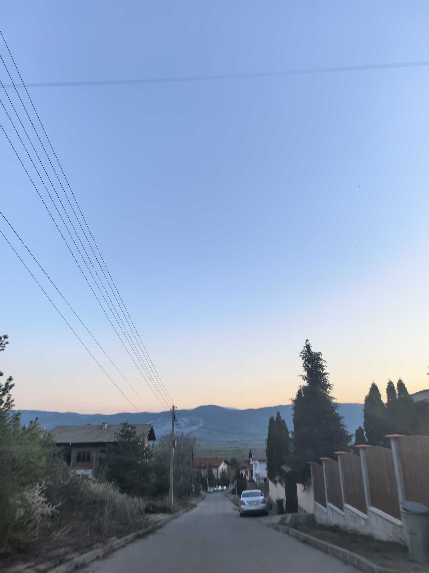 The image depicts a street in an unknown city during twilight. The sky is a gradient of light blue to orange near the horizon, with several power lines visible across the upper left portion. A road runs through the center of the frame, leading towards distant mountains. On either side of the road are houses and fences. A car is parked on the right side of the road. The environment includes trees, shrubs, and grass, suggesting a residential neighborhood. There is no visible interaction or explicit action occurring in the scene.