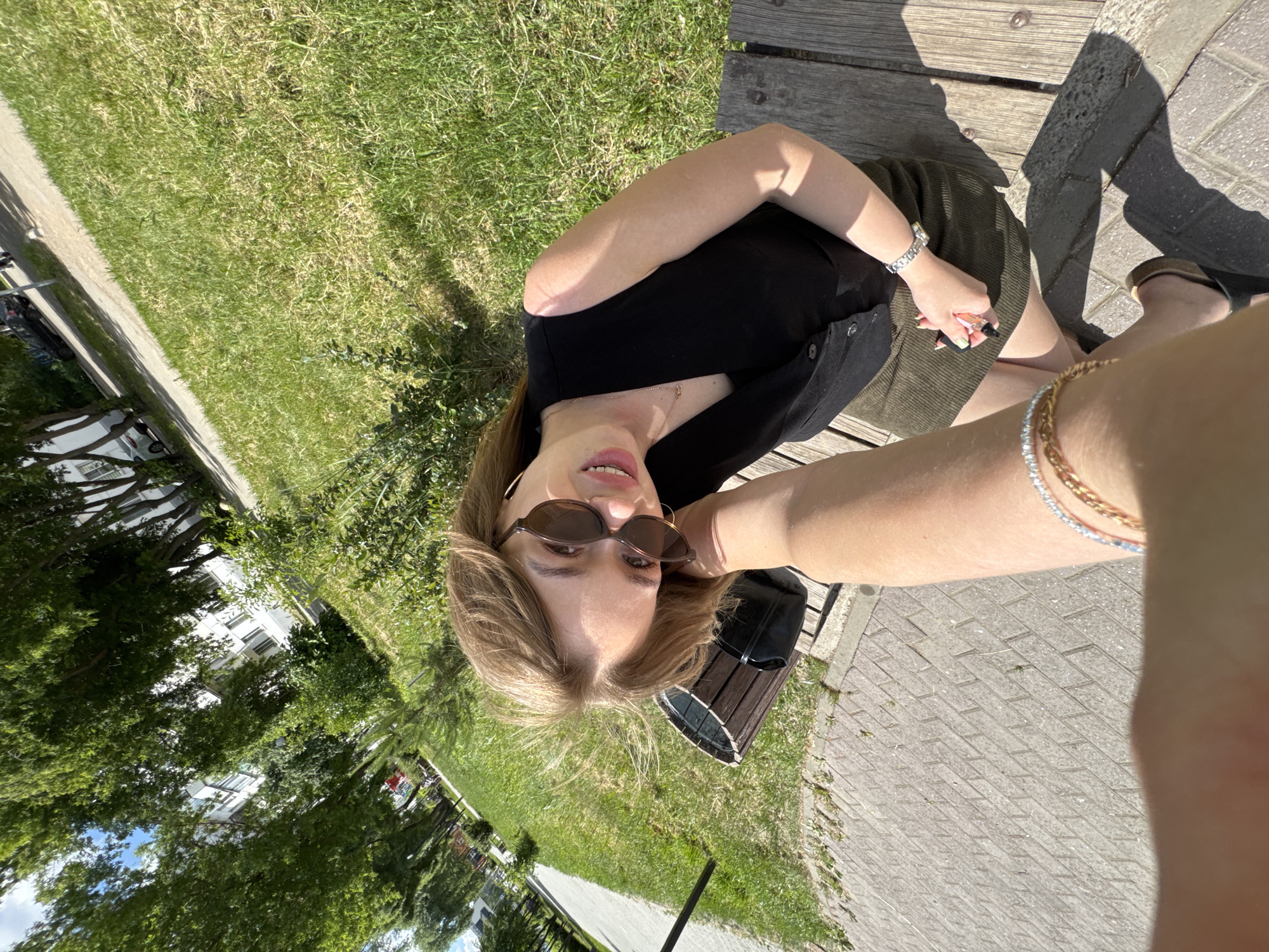 A young woman with blonde hair and sunglasses sits on a park bench. She is wearing a black vest over a dark top and a green corduroy skirt. She is smiling and looking at the camera. A person's arm, adorned with bracelets, is extended towards her. The background shows a paved path, green grass, trees, and a building. The scene appears to be a public park in the city of Chaikovsky, Russia, on a sunny day.
