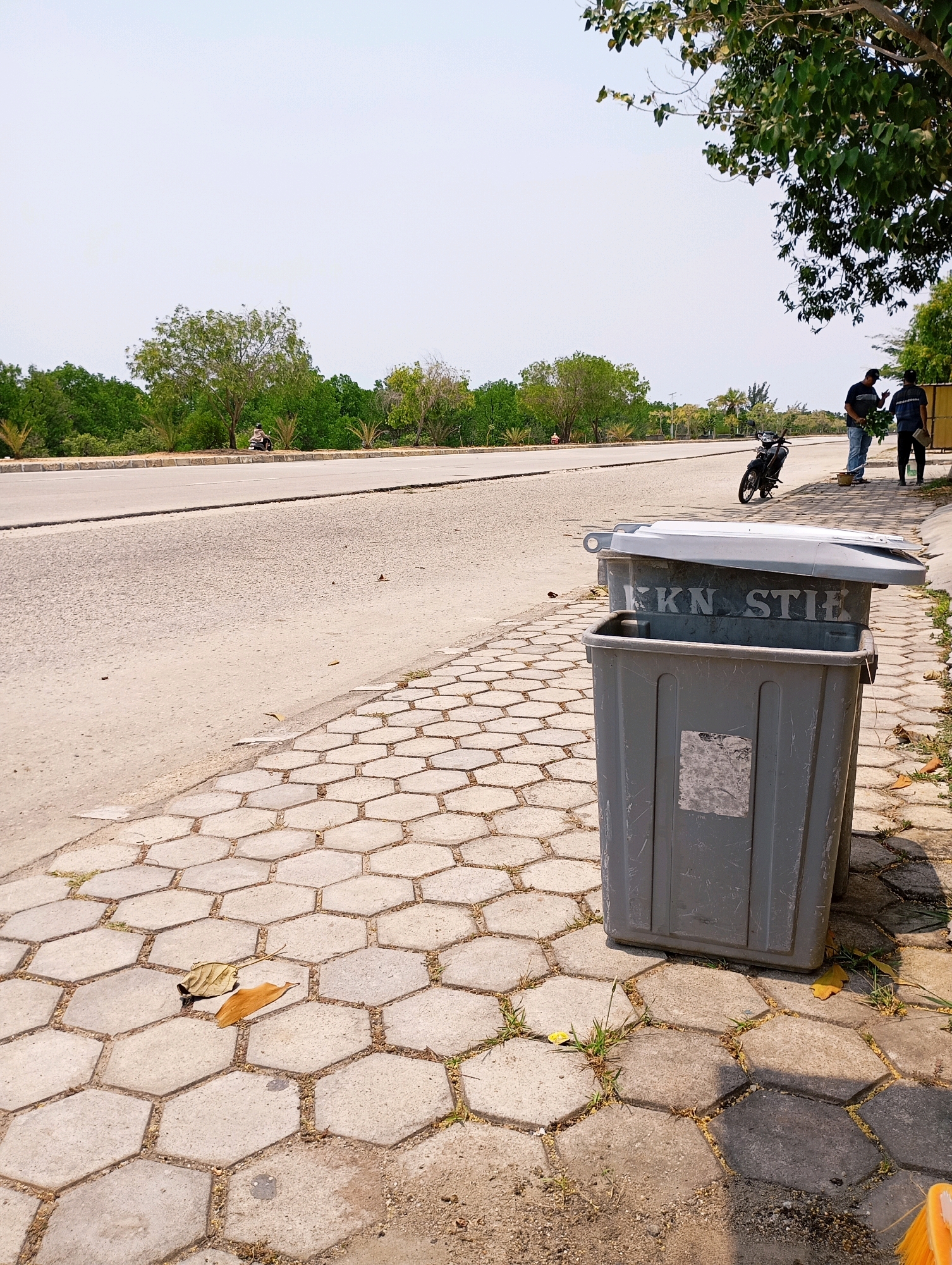 In Teluk Air, Indonesia, a gray trash can with a partially open lid stands on a hexagonal paved sidewalk next to a road. The word "FKKS TIE" is visible on the side of the trash can. A motorcycle is parked on the shoulder of the road. Two men stand near the motorcycle, one holding a bunch of green leaves. Trees and vegetation line the opposite side of the road. The sky is clear and bright.