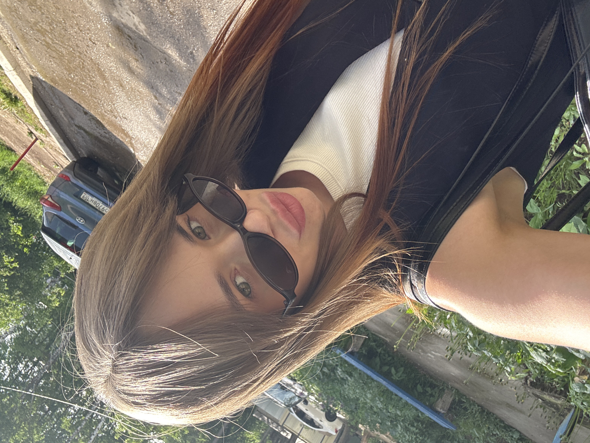 A woman with long brown hair is wearing sunglasses, a white turtleneck, and a black garment. She is outdoors in a setting that includes greenery, possibly a garden or park. In the background are cars and a structure which suggest an urban environment. The car has the license plate number "O970BH 159," indicating it is registered in the Perm Krai region of Russia. Chaikovsky is in Perm Krai.