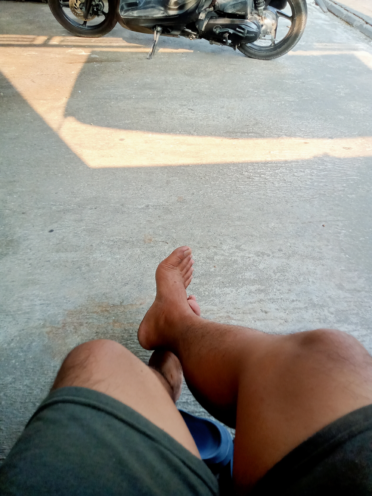The image shows a close-up of a person's bare feet and legs, with a motorcycle in the background. The ground appears to be concrete, with a shadow cast across it from an unseen object. The lighting suggests it is daytime, with the sun casting a warm glow. The overall impression is one of a relaxed, informal setting, possibly a residential area or a place where one might park a motorcycle. There is no indication of the specific location within Majan, Indonesia, beyond the general environment.