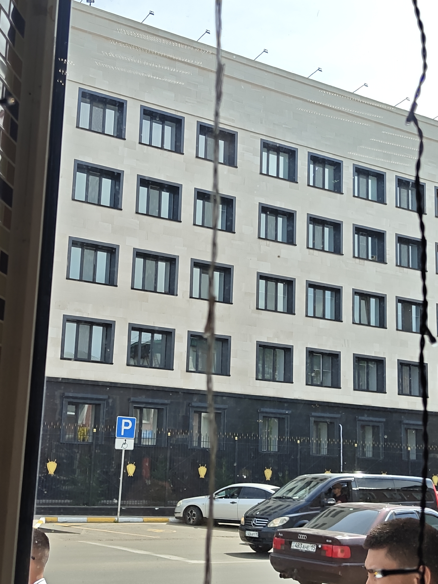 The image shows a modern, multi-story building with a light-colored facade and numerous dark-framed windows. The building appears to be an office or government building, possibly in the city of Petropavl, Kazakhstan. In front of the building is a black metal fence adorned with golden decorative elements. A parking sign with a blue "P" and a symbol for disabled parking is visible near the fence. Several cars are parked on the street in front of the building, including a silver sedan, a black minivan, and a burgundy sedan. A person is visible in the driver's seat of the black minivan. The sky above is bright, suggesting daytime. The image is framed by what appears to be a window or a similar viewing obstruction, with thin, dark vertical elements obscuring parts of the scene.