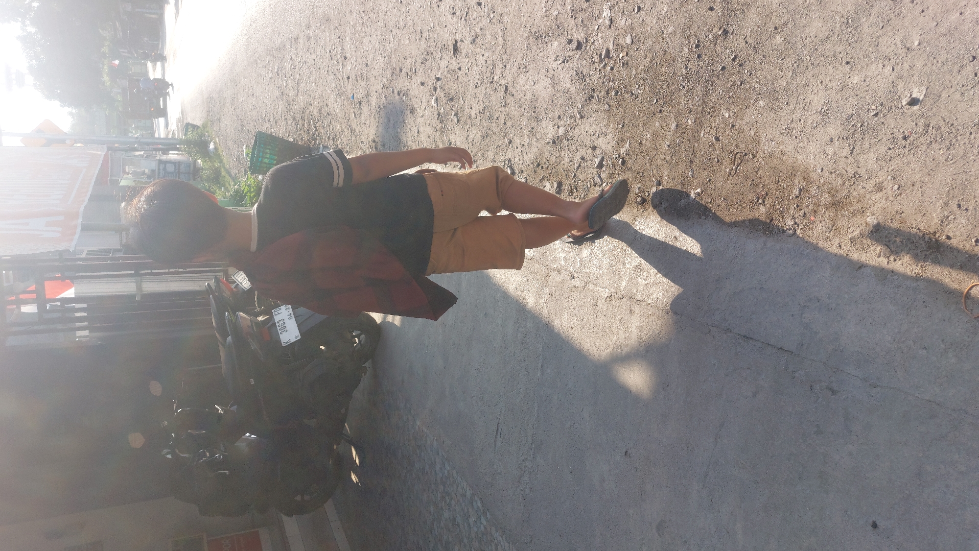 A man is walking on a paved path near a road in Karas, Indonesia. He is wearing a black t-shirt with white stripes on the sleeves, khaki shorts, and flip-flops. He is carrying a red and black plaid shirt draped over his left shoulder. To his left, a motorcycle is parked. The scene appears to be outdoors during the daytime, with sunlight casting a shadow of the man on the ground. The ground is a mixture of dirt and pavement, with some gravel and small stones. In the background, there is a blurry view of buildings and vegetation. The overall atmosphere is casual and relaxed. There is no visible text in the image.