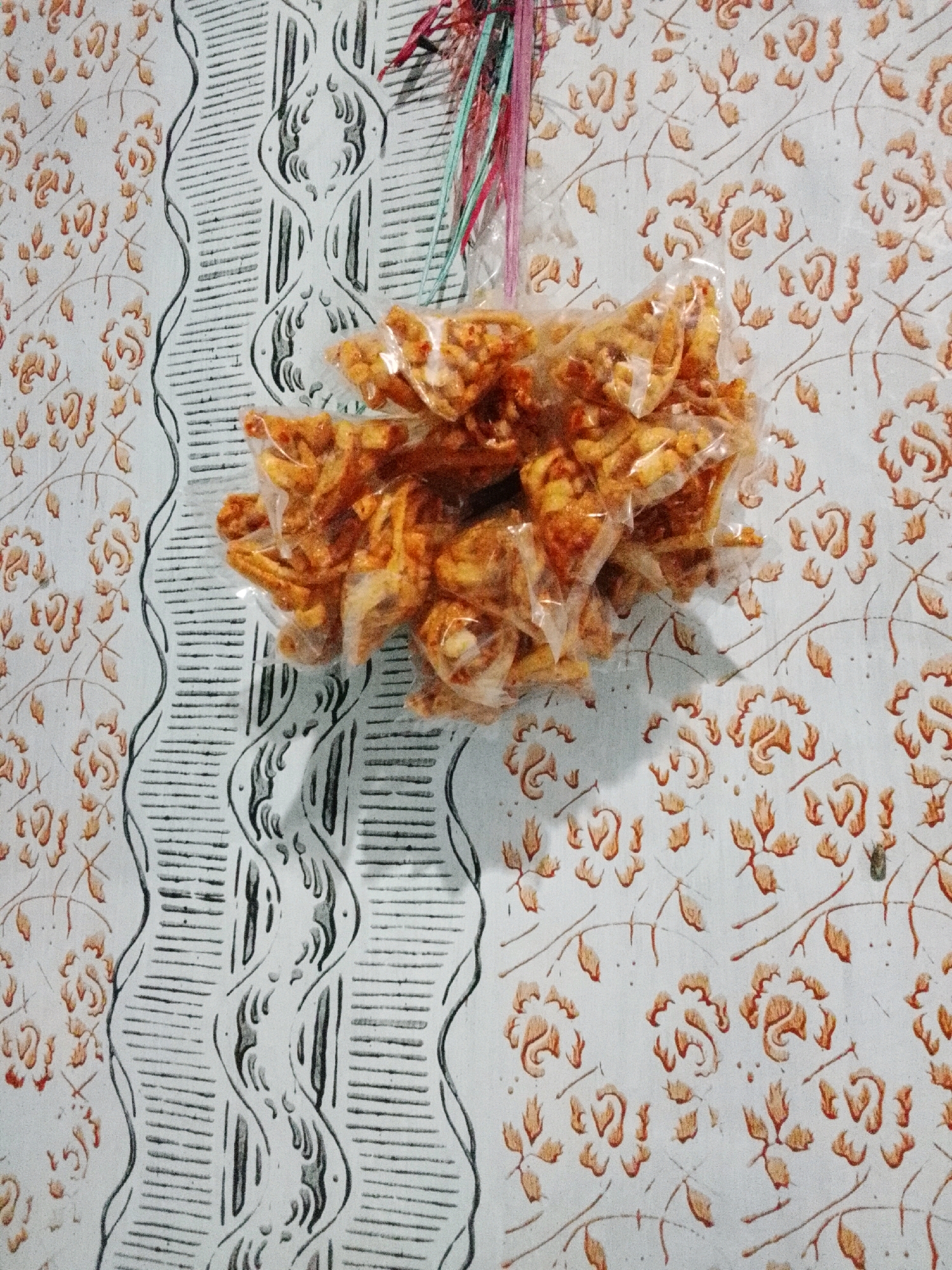 A bag of crispy snacks is hanging by colorful strings against a patterned wall. The snacks are golden brown and appear to be seasoned with spices. The wall features a repeating floral motif in earthy tones and a vertical stripe pattern with wavy lines and horizontal striations. The scene is an indoor setting, likely in a home. The specific location of Padang Dalom, Indonesia, is not discernible from the visual information provided in the image.