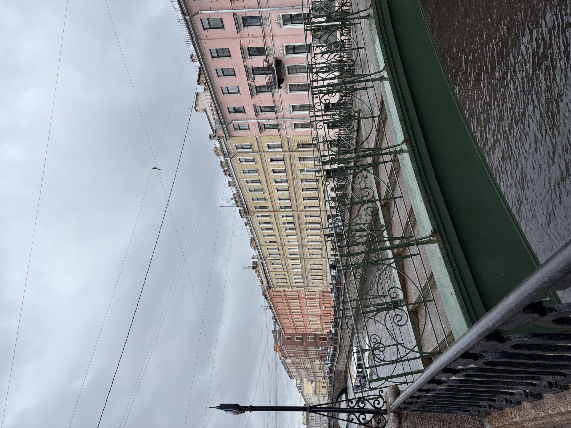 The image shows a street scene in Saint Petersburg, Russia, taken from a bridge. The perspective is angled, with a row of colorful buildings on the left side of the frame. These buildings are painted in shades of pink, yellow, and red, and they appear to be apartment buildings. The architecture suggests a historic European style.
In the center of the frame, there's a canal or river. The water's surface is dark and reflects the overcast sky. A green-painted edge of the bridge is visible, adding to the architectural structure.
The railing of the bridge is made of decorative wrought iron. The sky is overcast, suggesting a cloudy or rainy day. A street lamp is visible in the lower left corner of the frame. Power lines stretch across the sky. There are cars parked along the street.