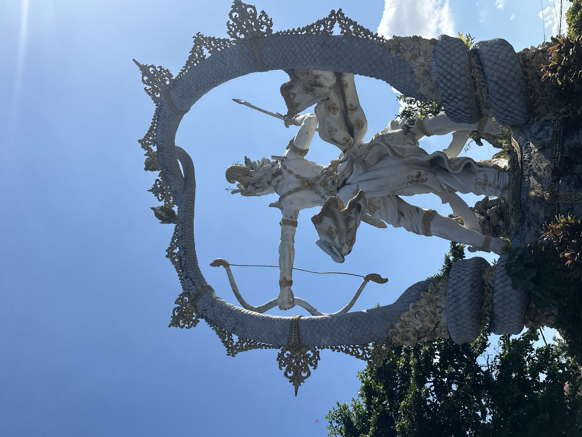 This image shows the Arjuna statue in the city of Ubud District, Indonesia. The statue depicts Arjuna, a central character from the Hindu epic Mahabharata, in a dynamic pose with a bow and arrow. He is made of white stone and adorned with gold accents. The statue is framed by a large, circular archway resembling a serpent, with intricate decorative elements. The sky is a clear blue with some white clouds, and lush green trees are visible on the left side of the frame. The sun is shining brightly, casting a slight glare.