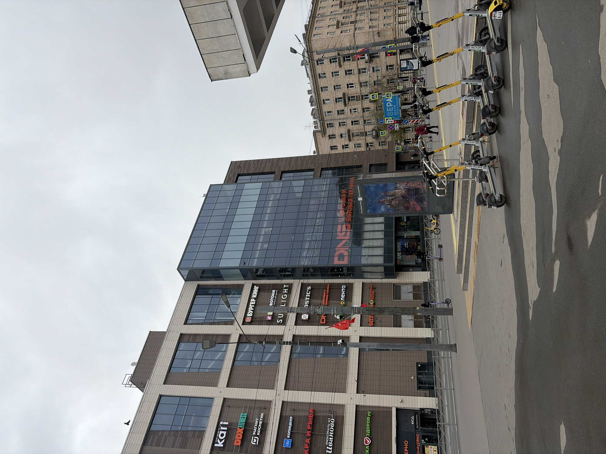 This image captures a vibrant urban street scene in Saint Petersburg, Russia, under an overcast sky. The photograph is taken from a slightly low angle, looking up at two prominent buildings and a street area.
In the foreground, a row of bright yellow and grey electric scooters, branded "WHOOSH," are neatly parked on what appears to be a sidewalk or designated scooter parking area. Several scooters display identification numbers such as "AF4758," "AP3710," "AN6108," and "HK6 84E." The ground features asphalt pavement with visible white and yellow pedestrian crossing markings, and long shadows stretch across the surface.
To the left, a modern multi-story commercial building dominates the frame. Its facade is a mix of large, reflective glass windows and textured brown or tan panels. Numerous store signs with Russian text are visible, including "kari," "DDXFITNESS," "МАГНИТ КОСМЕТИК" (Magnit Cosmetik), "БУРГЕР КИНГ" (Burger King), "ВОТОНЯ," "SU LIGHT," "КОПИЦЕНТР" (Copy Center), "КРА И РЫБА," "текстиль из Иваново" (Textiles from Ivanovo), "ЕКА ЗДОРОВ ру," "SHAVERNO," and "ВХОД" (Entrance). Higher up, larger signs read "DN" (likely DNS, a tech retailer) and "OSTIC'S." A large billboard advertises "ЦИФРОВАЯ И БЫТОВАЯ ТЕХНИКА" (Digital and Home Appliances).
In the background, behind the modern building, stands an older, more traditional residential building with numerous windows, suggesting a typical city block. Various street furniture, including a tall lamppost, traffic lights, and multiple road signs, are scattered throughout this area. Notable signs include pedestrian crossing indicators and a blue sign with "BЕРА 92.9 FM" (Vera 92.9 FM), indicating a radio station. A few blurry figures of people can be seen walking in the distance, engaging in everyday urban activity. The overall impression is one of a bustling city environment on a cloudy day.
