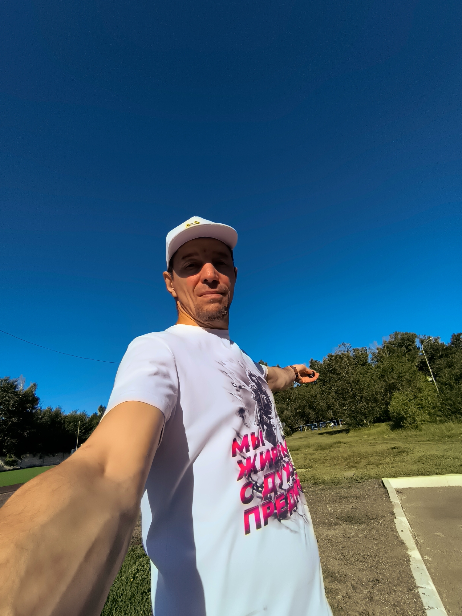 A man wearing a white t-shirt and a white baseball cap is standing outdoors on a bright, sunny day, with a clear blue sky above. He is pointing to his right with his left hand, holding a small, dark object. The man has a goatee and appears to be in his late 30s or early 40s. His t-shirt has a graphic design with pink lettering that reads "МЫ ЖИВЕМ С ДУШОЙ" in Russian, which translates to "WE LIVE WITH SOUL". The background consists of a grassy area, trees, and a paved surface. The man's arm is extended towards the viewer, suggesting he is taking a selfie or being filmed. The scene is outdoors, and there is no visible indication of the specific city or location.