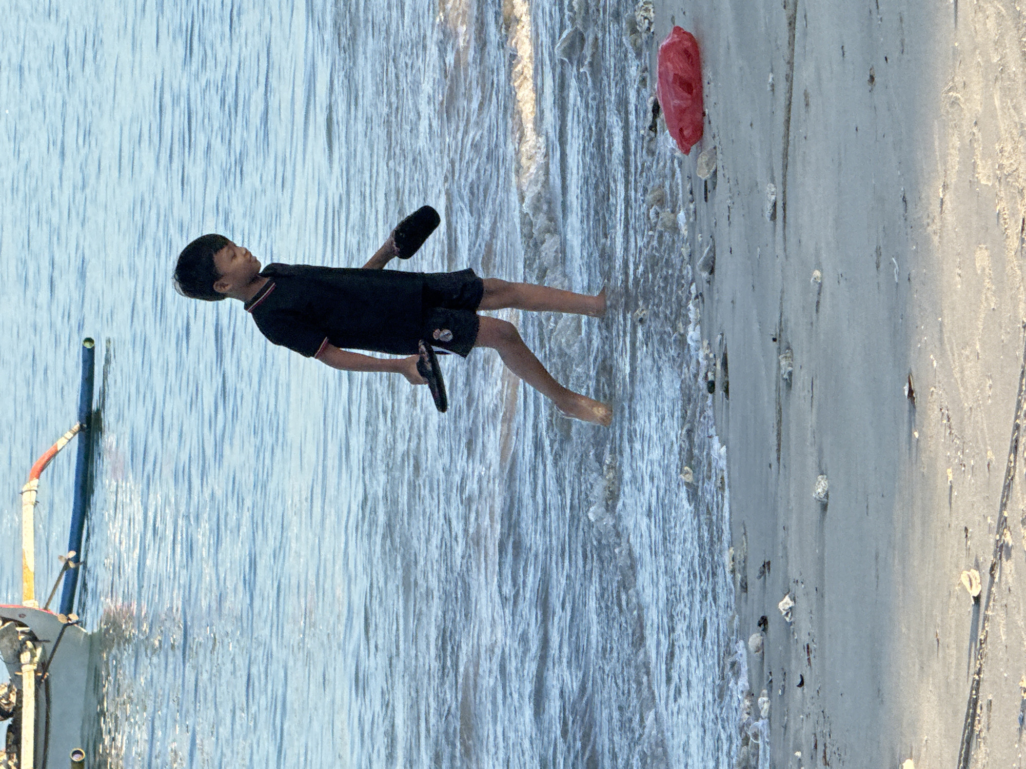 A young boy in a black t-shirt and shorts walks along the shore at Kuta Beach, Bali, Indonesia. He is holding a pair of black flip-flops in his hands. The water is shallow and calm, with small waves lapping at the sand. The beach is littered with small stones and coral fragments. A red plastic bag is visible on the sand in the lower right corner of the frame. In the background, a white boat with outriggers is anchored in the water. The sky is overcast, and the light is soft.