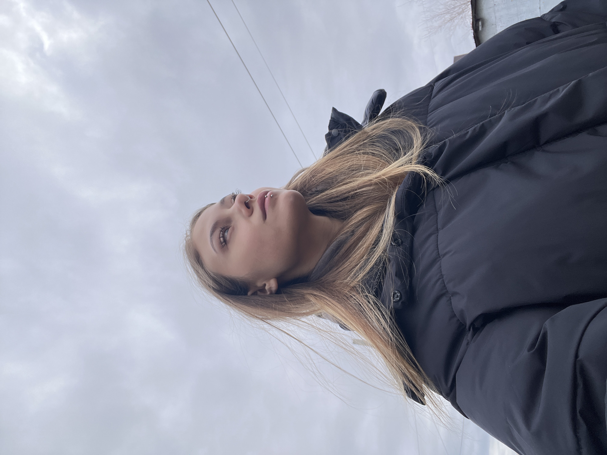 A low-angle shot of a young woman with light brown hair wearing a black puffer jacket. Her head is tilted back, looking upwards. She has a piercing on her upper lip. The background features a cloudy sky, with power lines visible in the upper left corner. A building's edge is seen in the upper right corner. It is not possible to confirm the location of Фоки, Russia from the image.