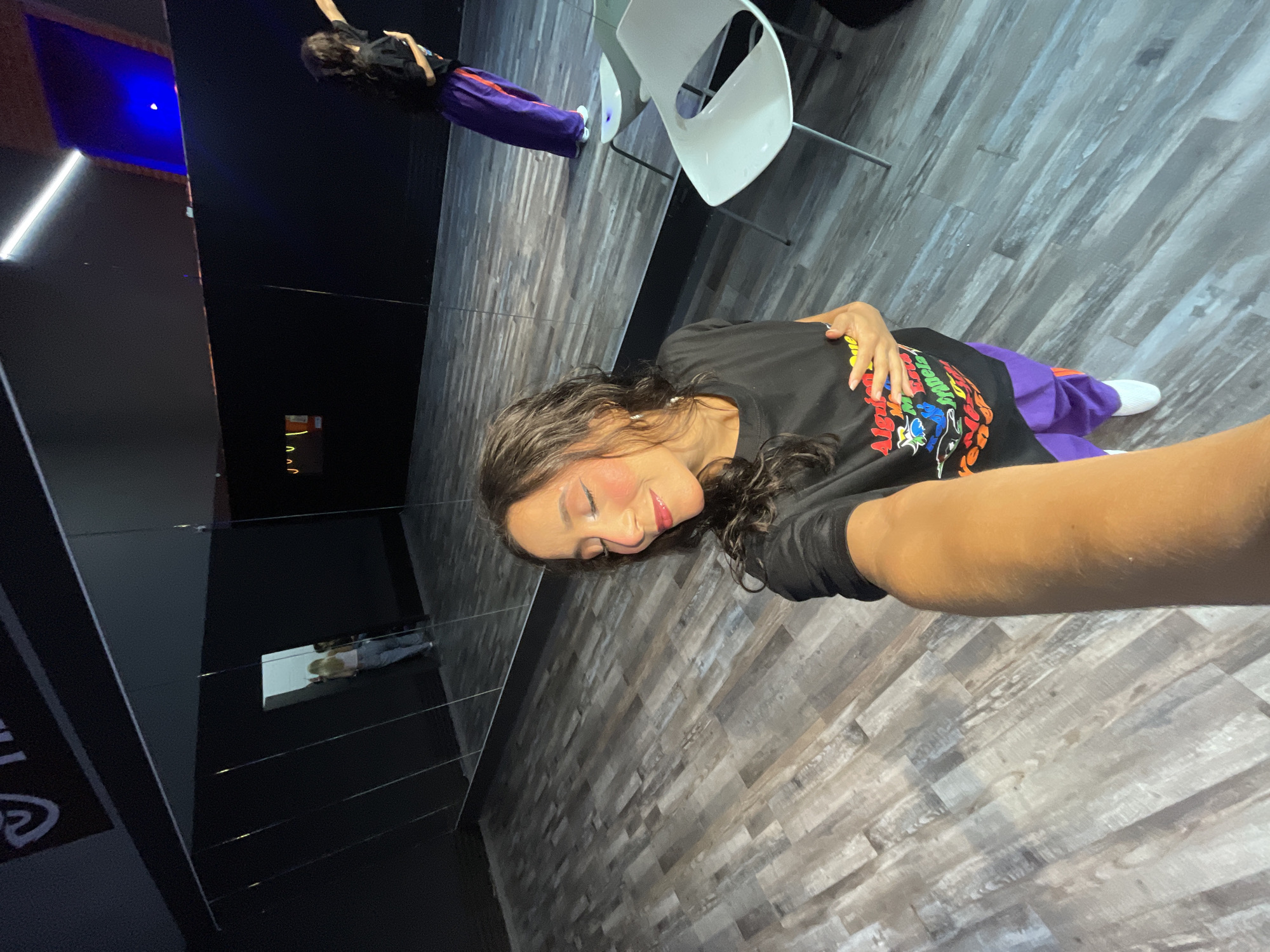 In a dance studio in Pampatar, Venezuela, a woman of Latin American descent, with dark wavy hair and fair skin, is taking a selfie. She is wearing a black t-shirt with a colorful graphic design and vibrant purple pants. Her eyes are closed and she has a smile on her face, with heavy pink blush and red lipstick. The studio has a dark-colored wall with large mirrors reflecting the space. The floor is made of wooden planks. In the reflection, another woman with blonde hair is visible near a doorway, and a second person in a black t-shirt and purple pants is standing further away, facing a mirror. Two white chairs are positioned in the foreground, near the woman taking the selfie.