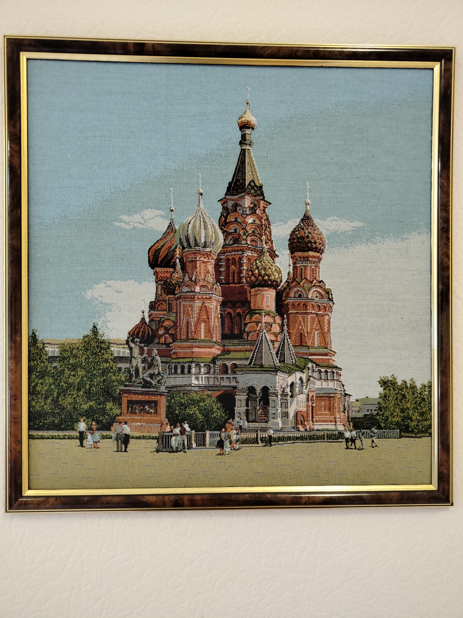 The image is a framed tapestry depicting Saint Basil's Cathedral in Moscow, Russia. The tapestry shows the iconic onion-domed cathedral with its vibrant red and multi-colored domes. In front of the cathedral, there is a square with a few people walking and standing around, as well as a horse-drawn carriage with two people. Trees and other buildings are visible in the background. The sky is light blue with some white clouds. The tapestry is framed in a dark wood frame with a gold inner trim. The location of Березово (село) (новокузнецкий), Russia, is irrelevant to the content of the image, which clearly depicts a famous Moscow landmark.