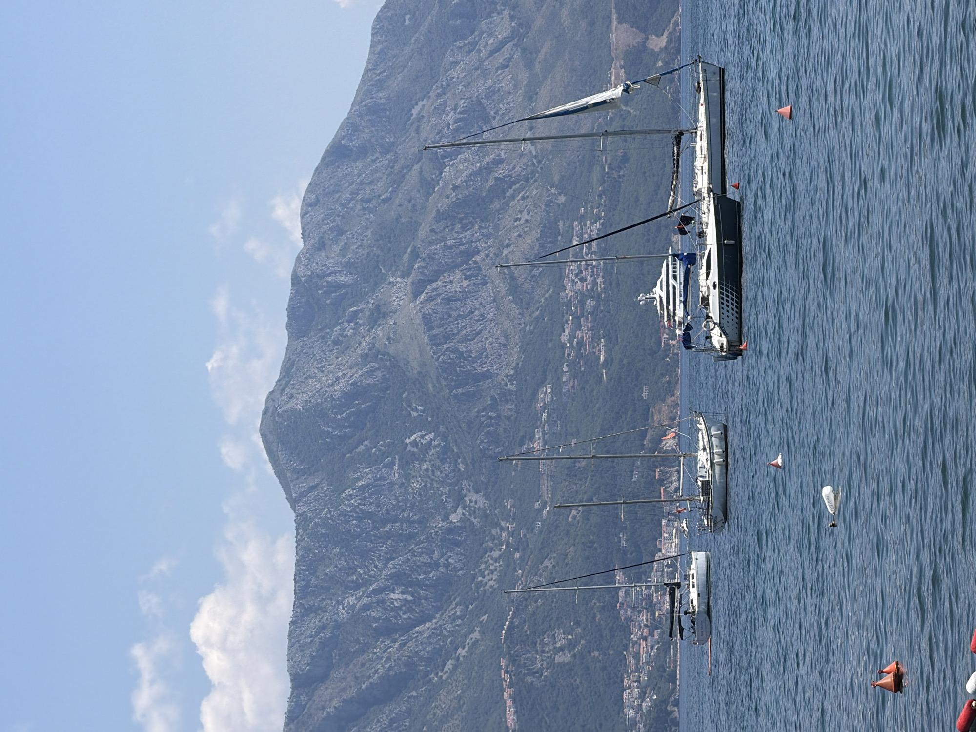 The image displays a fleet of sailboats on a calm, blue body of water. The boats are arranged in a line, with their masts and sails prominent against a backdrop of a steep, rocky mountain covered in greenery. Buildings are visible clustered on the mountainside, suggesting a coastal town or village. The sky is bright blue with a few scattered white clouds, indicating fair weather. The lighting suggests it is daytime, likely late morning or early afternoon. The water has a textured surface with ripples, suggesting a gentle breeze. There are several buoys floating on the water, some orange and some white. One of the white buoys appears to have some text on it, but it is not clearly legible. The scene evokes a sense of tranquility and leisure associated with sailing. There are no discernible people in the image.