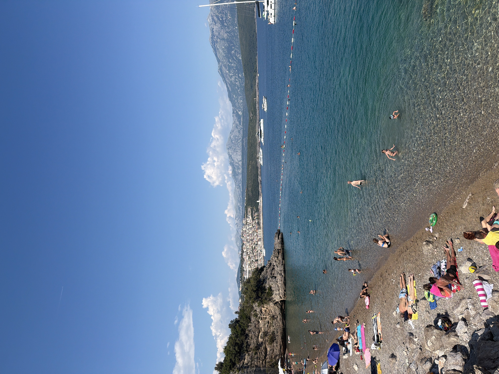 A high-angle view shows a crowded pebble beach in Radovići, Montenegro, bordering a clear, turquoise sea. Numerous people are scattered across the beach, some sunbathing on towels and blankets, others wading or swimming in the water. A few children are playing near the shore, and some are in deeper water. The sea is dotted with several boats, including a large white sailboat on the right and several motor yachts further out. A floating rope barrier marks a swimming area in the sea. In the background, a densely populated coastal town with white buildings rises up a hill, backed by lush green slopes and rocky mountains under a bright blue sky with scattered white clouds. The overall scene depicts a lively summer day at a popular beach destination.